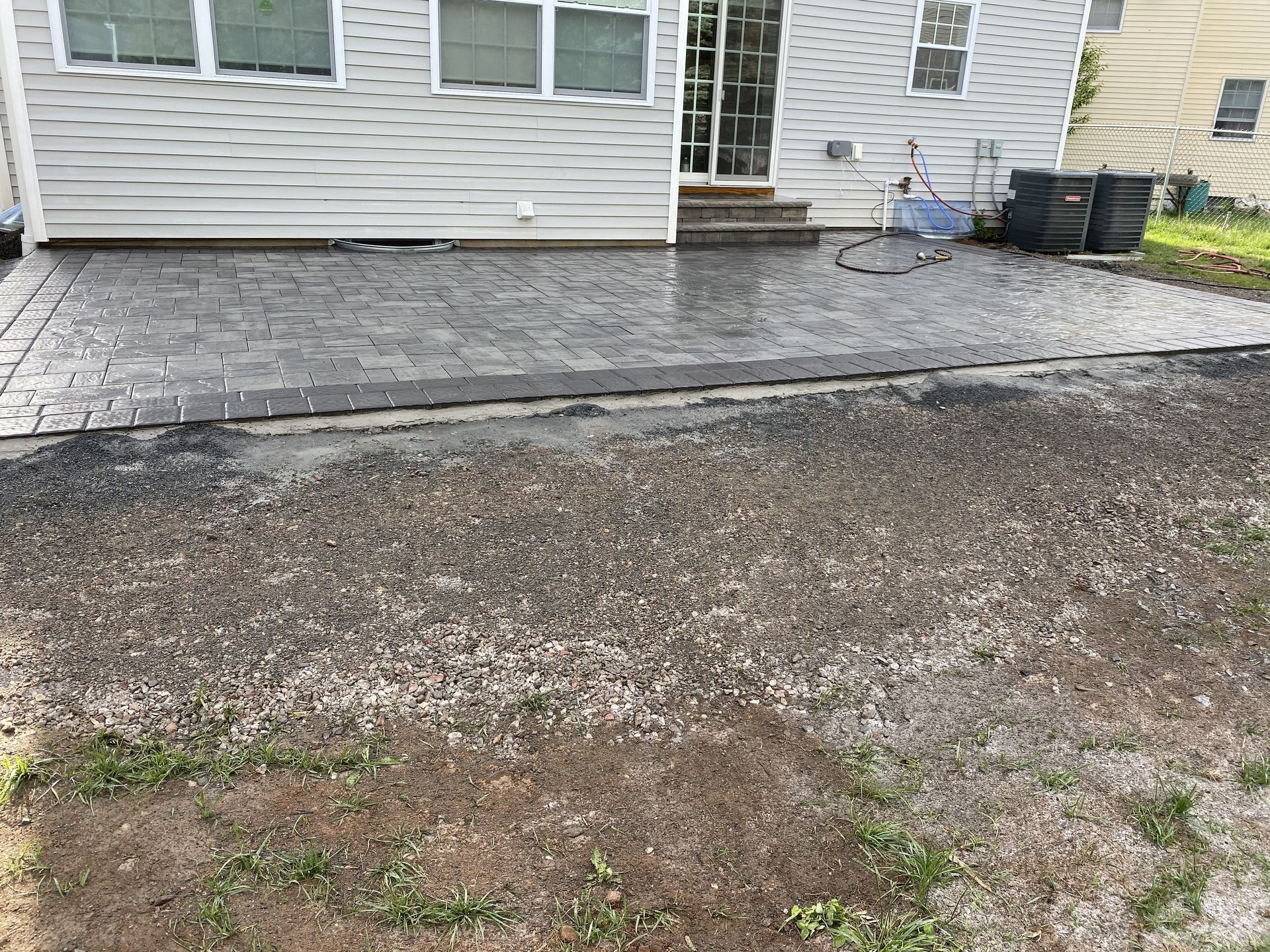 A newly installed gray stamped concrete patio sits against the back of a house, transitioning to a gravel yard.