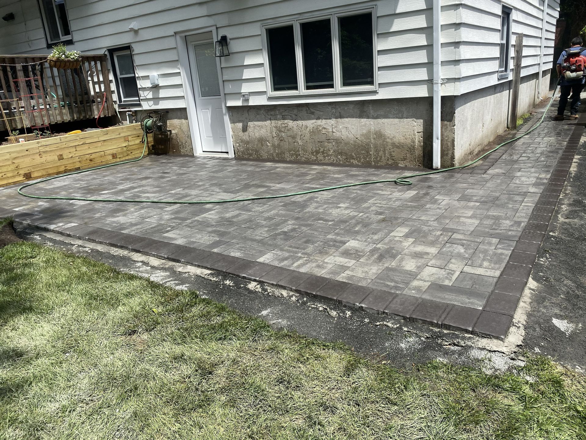 A newly installed gray paver patio with a dark border sits next to a white house, wooden steps, and a grassy lawn.