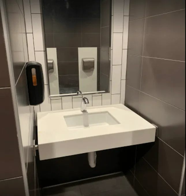 A modern public restroom sink, with a white countertop and dark gray tiled walls.