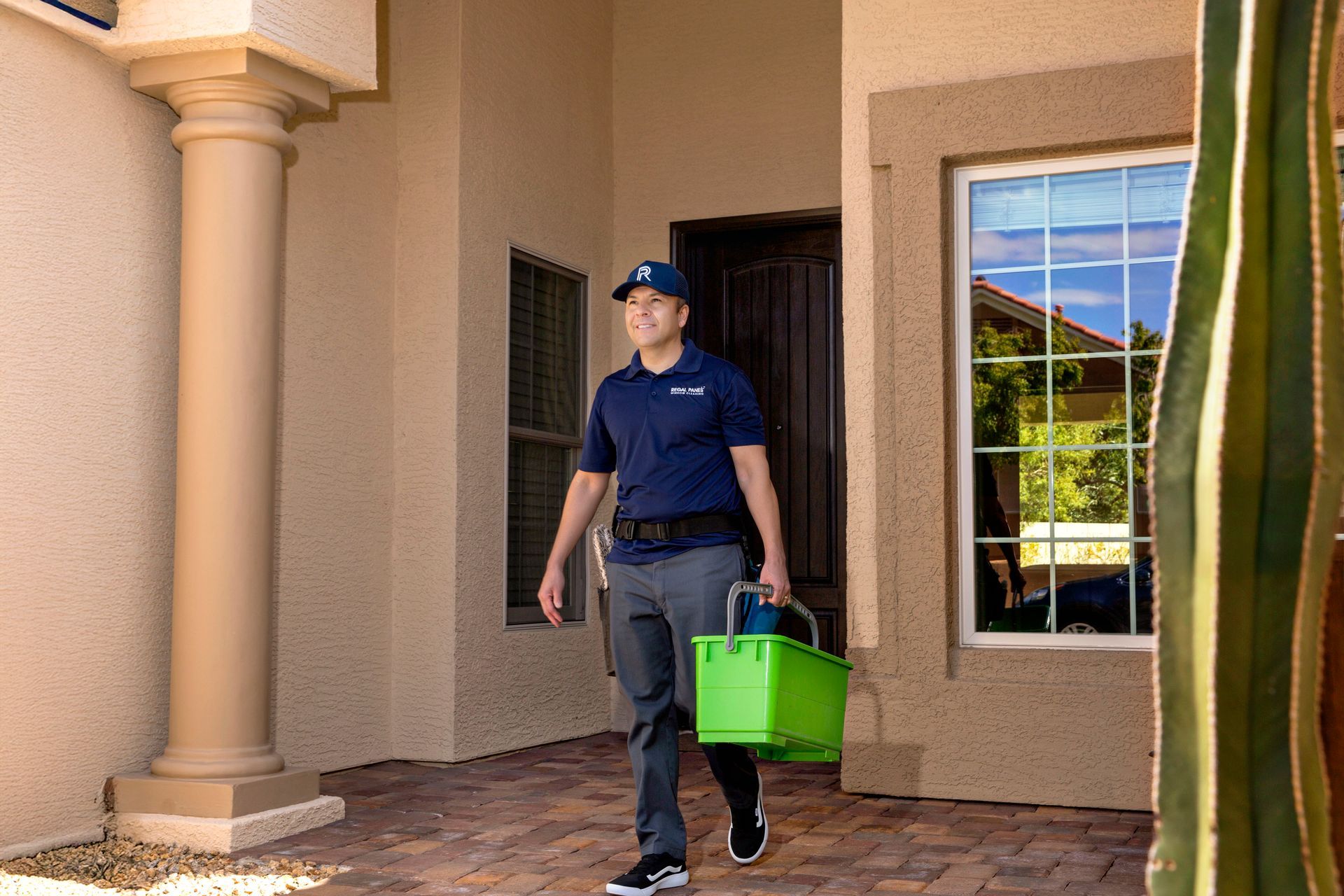 Person carrying a green bucket walks on a porch beside a window and columns.