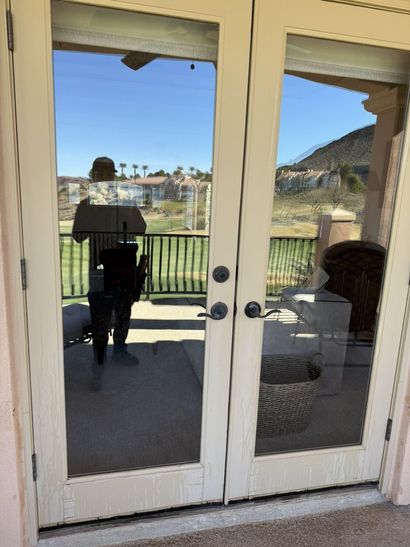 Glass patio doors reflecting a person standing on a porch, with a grassy yard and blue sky beyond.