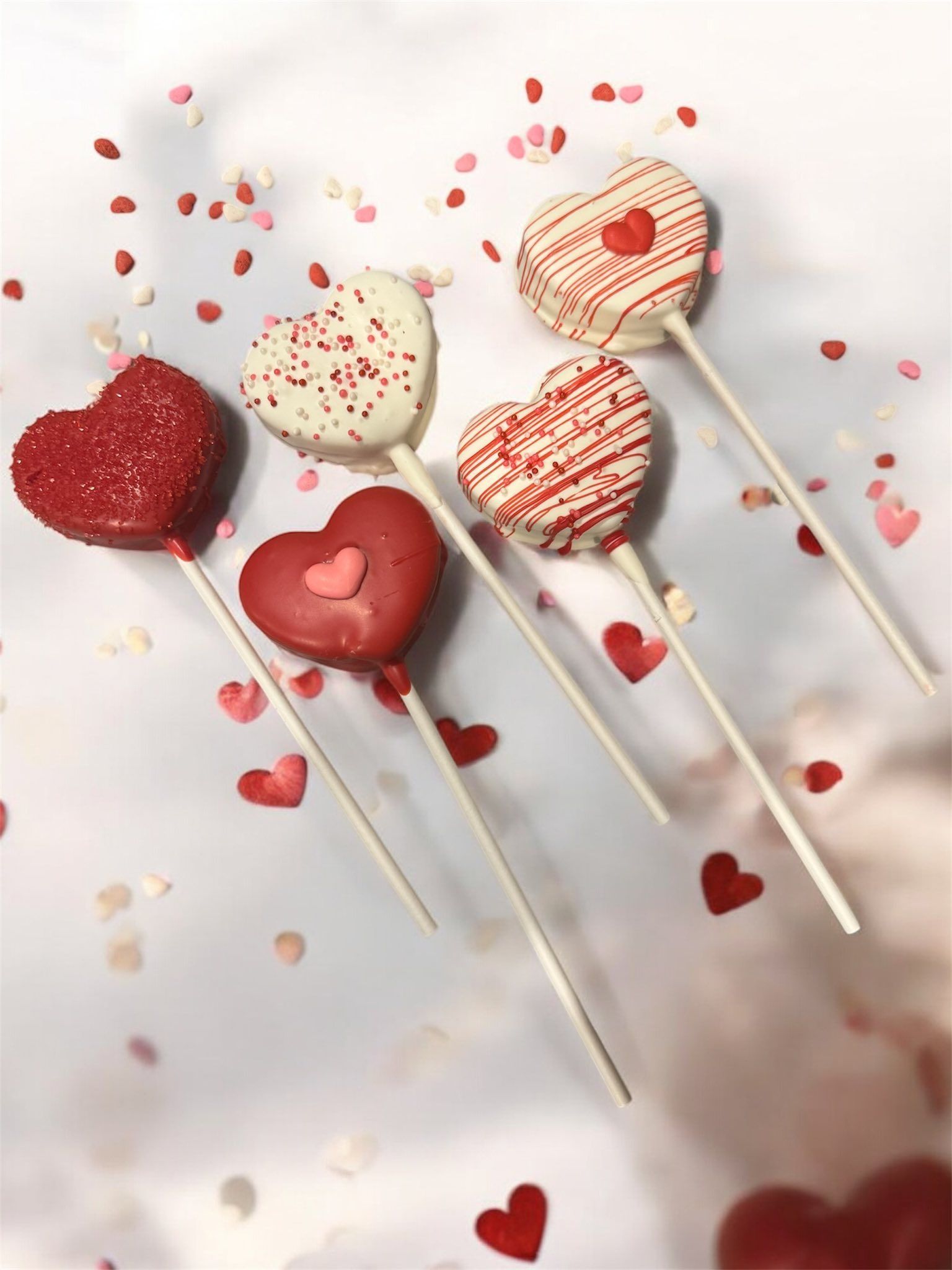 Heart-shaped decorated cookies on sticks scattered with red heart confetti on a light background