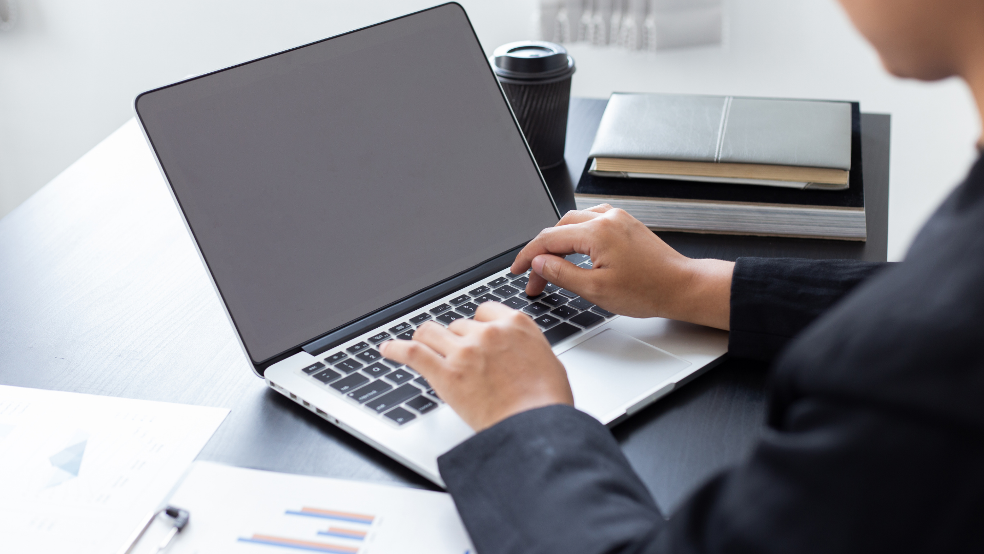 Person typing on a laptop at a desk with a coffee cup and notebook nearby