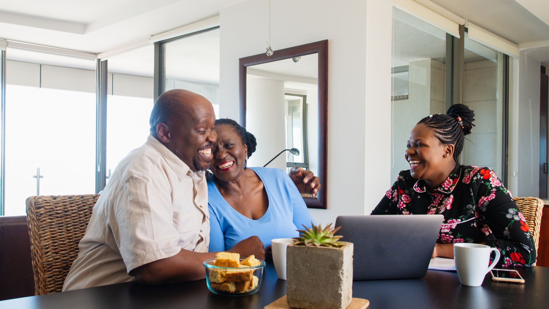 Three people smiling at a dining table with laptops and coffee in a bright home living room.