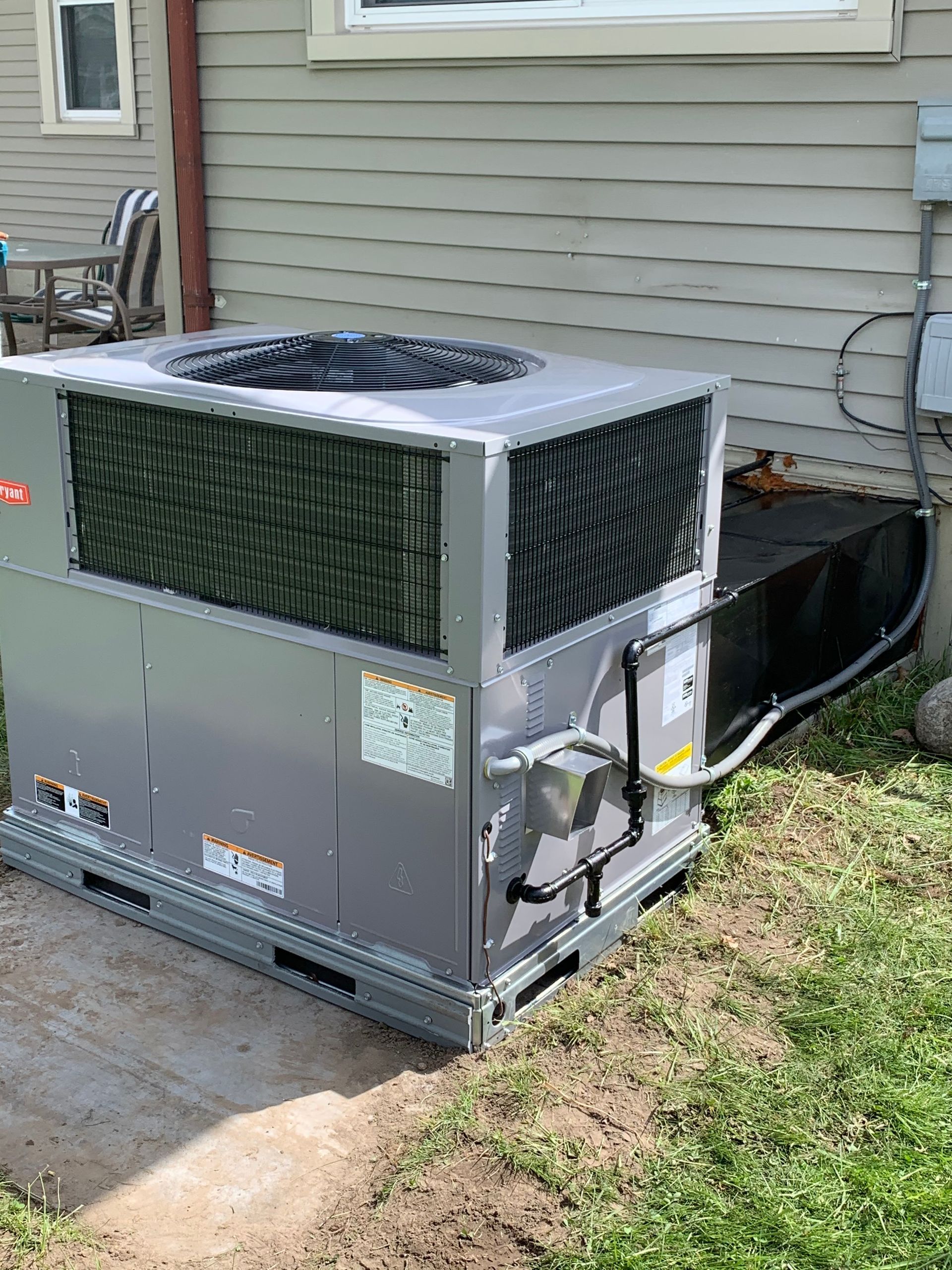 Gray HVAC unit next to a light gray house. Unit is on a concrete pad with a black metal duct.