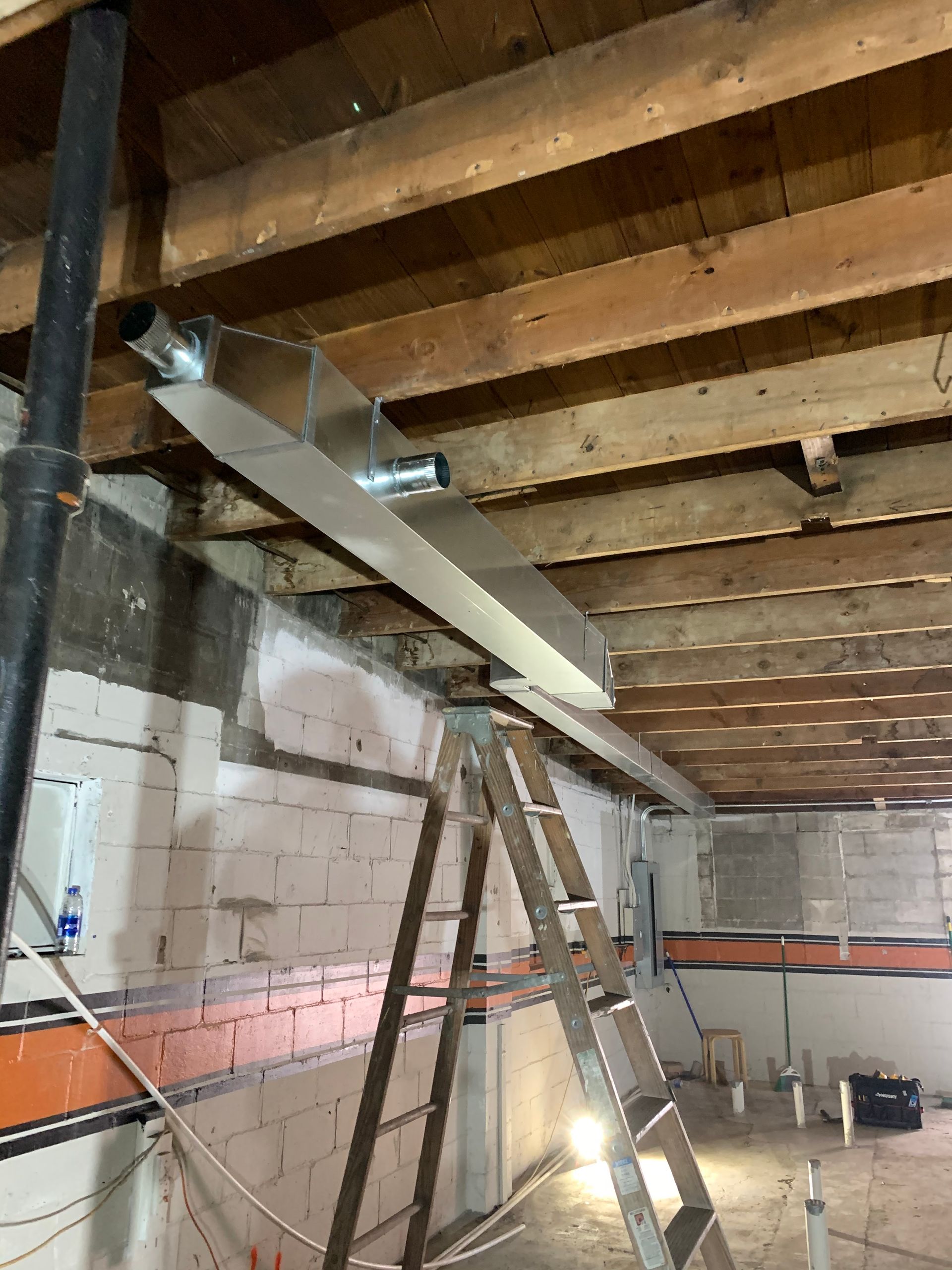 Construction site with metal ductwork installation. Ladder, wood beams, and concrete walls are visible.