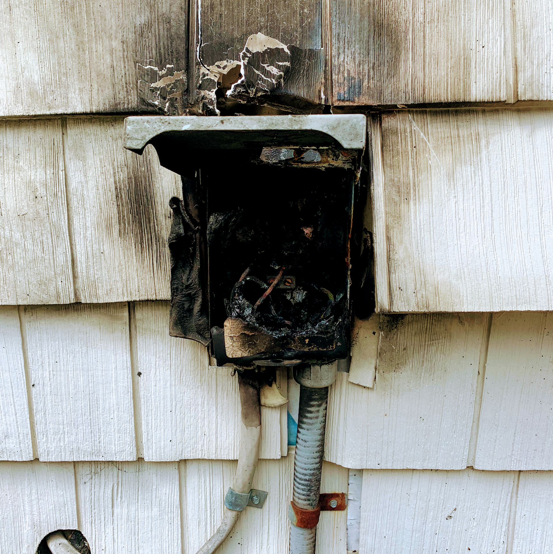 Burnt electrical box on a white shingled wall with charred debris and exposed wires.