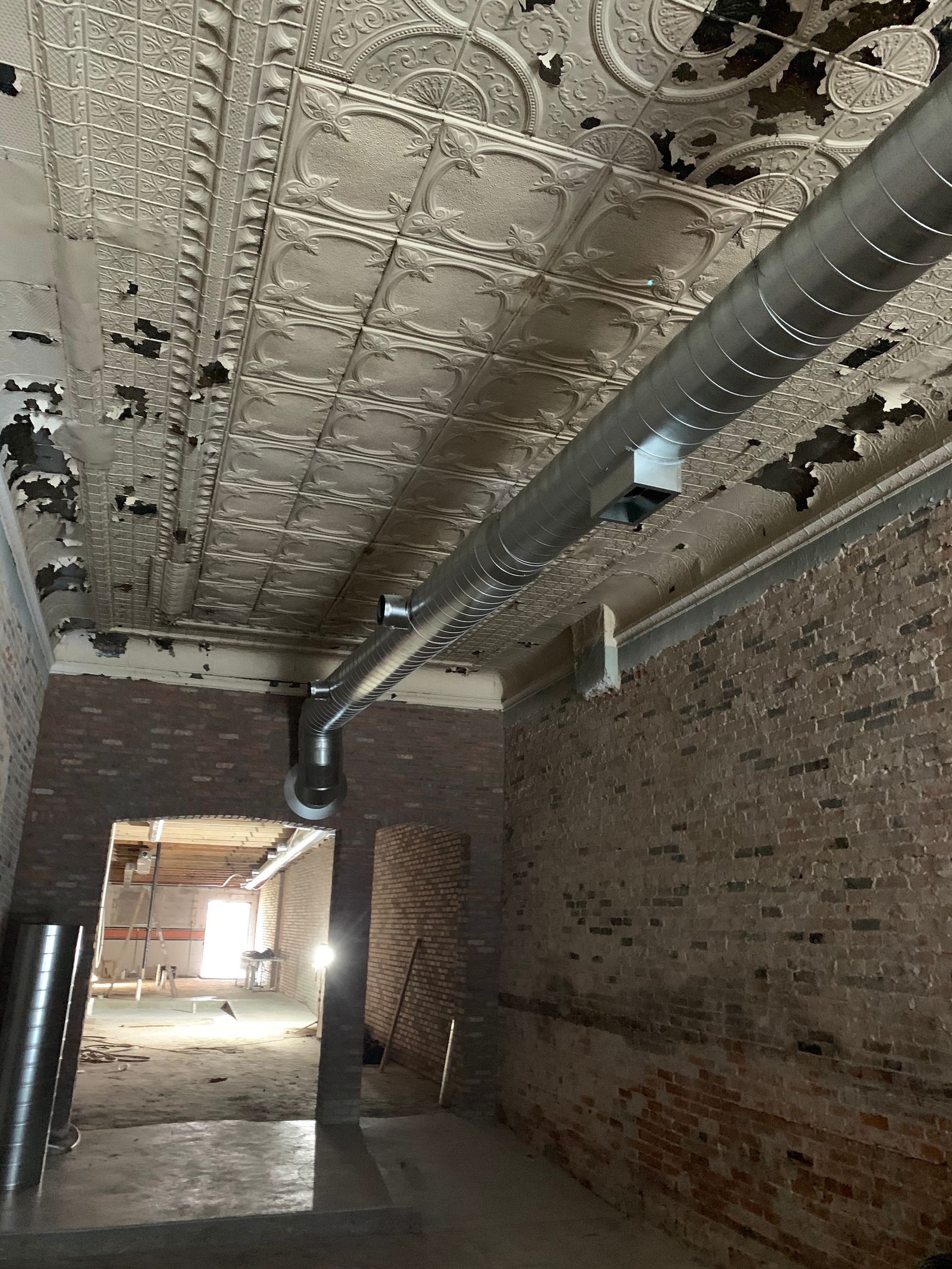 An interior view of a room with exposed brick walls, ornate peeling ceiling, and HVAC ductwork.