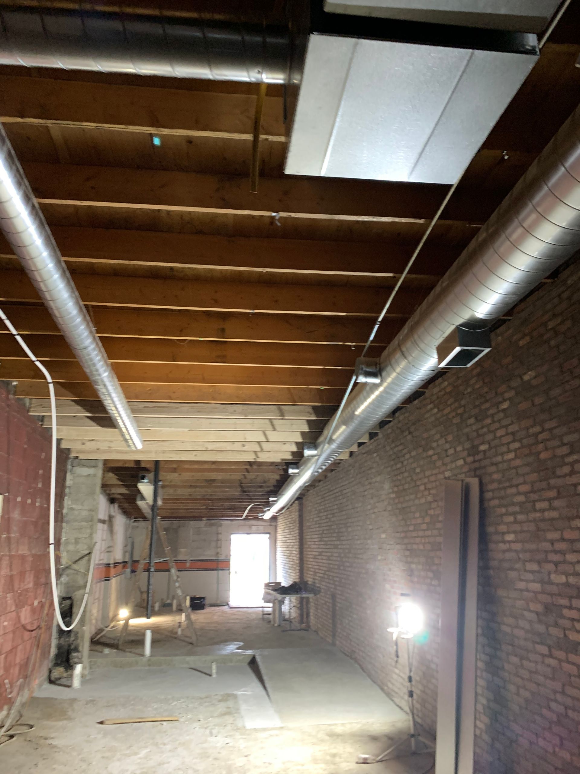 Interior of a room under construction with exposed brick wall, ductwork, and open doorway at the far end.