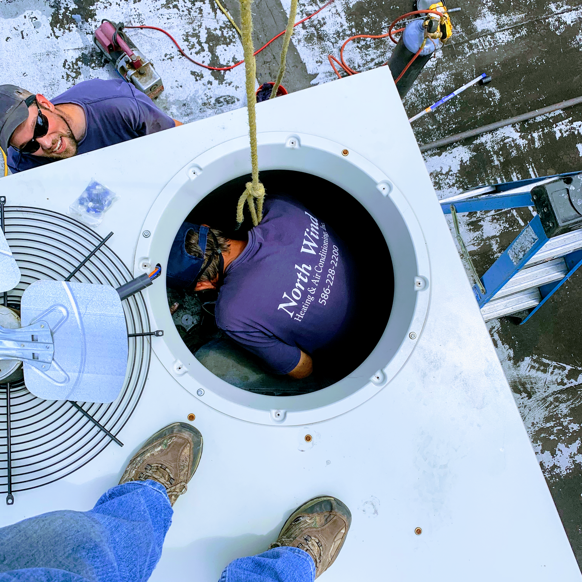 Two HVAC technicians working on rooftop equipment. One in equipment, one outside, both in blue.