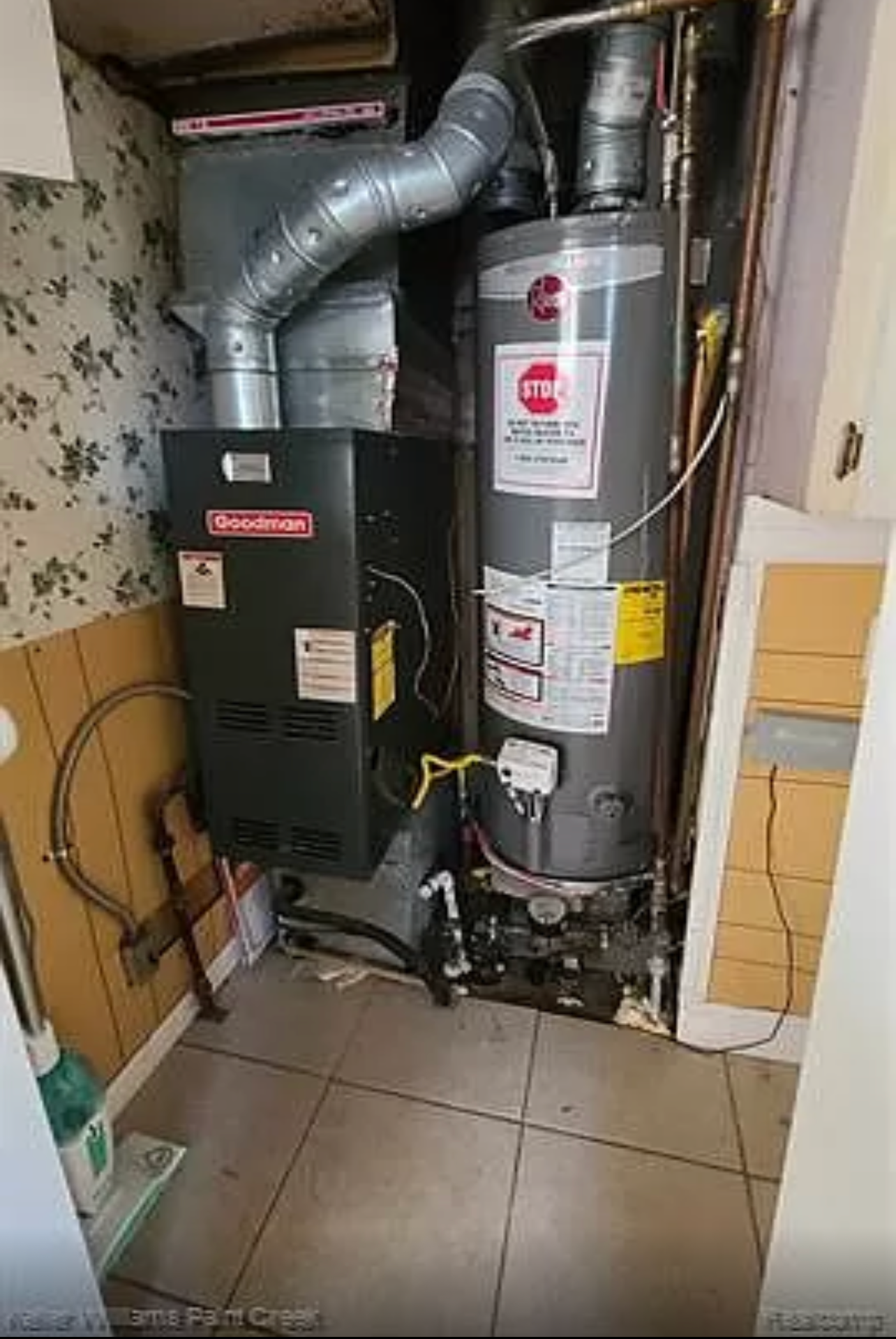 A small room with a furnace, water heater, and connecting pipes. Floral wallpaper on the walls.