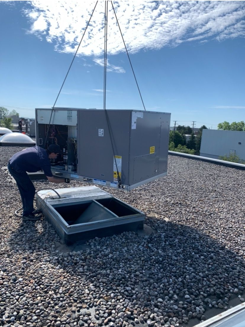 HVAC unit being lifted onto a rooftop by a crane. Workers are present, and the sky is blue.