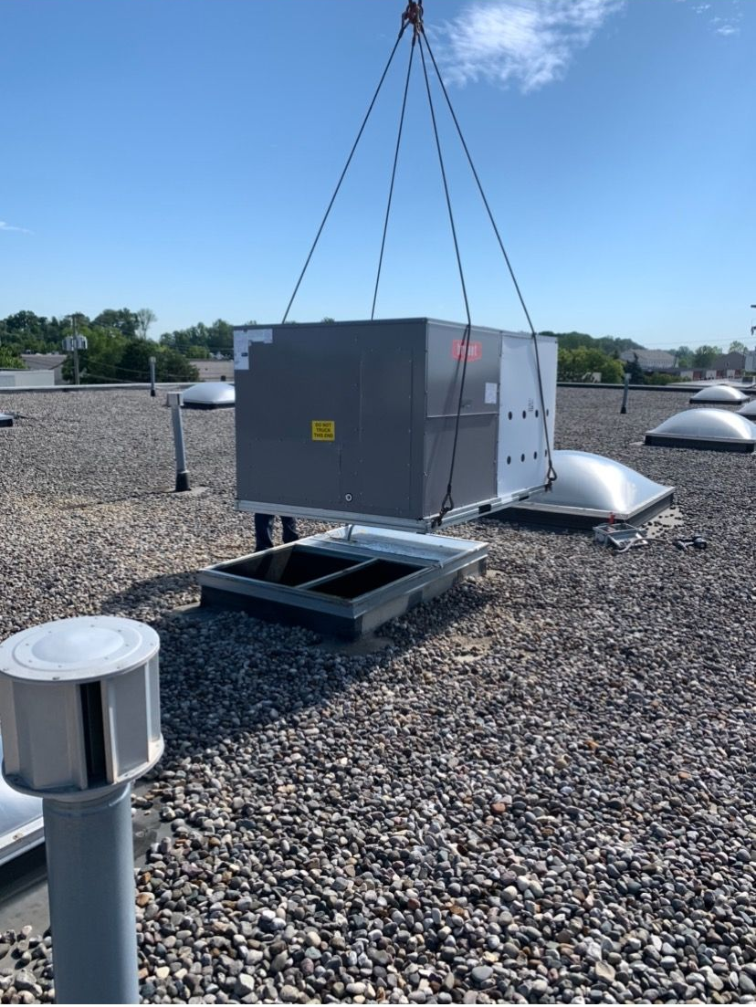 HVAC unit being lifted by crane onto a gravel rooftop on a sunny day.