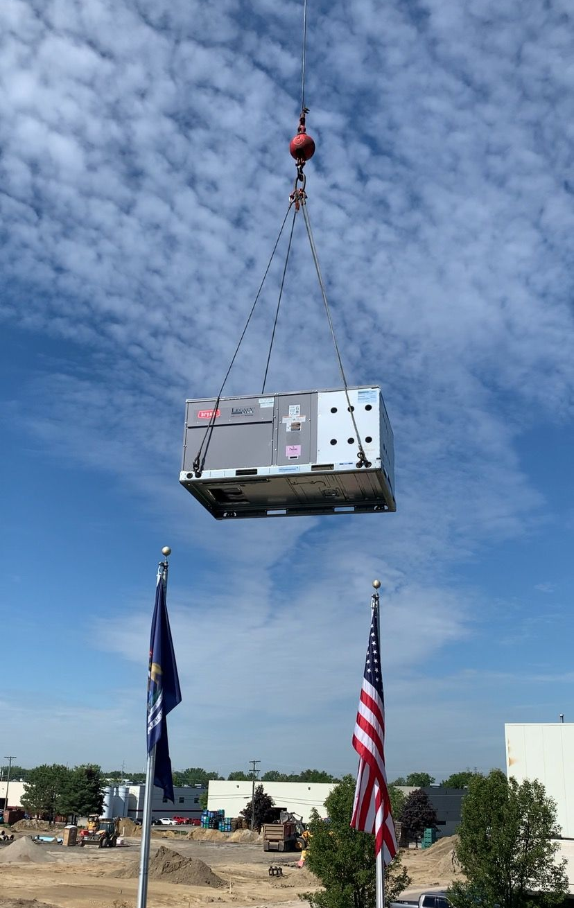 A large metal box is lifted by a crane against a partly cloudy blue sky. Flags are in the foreground.
