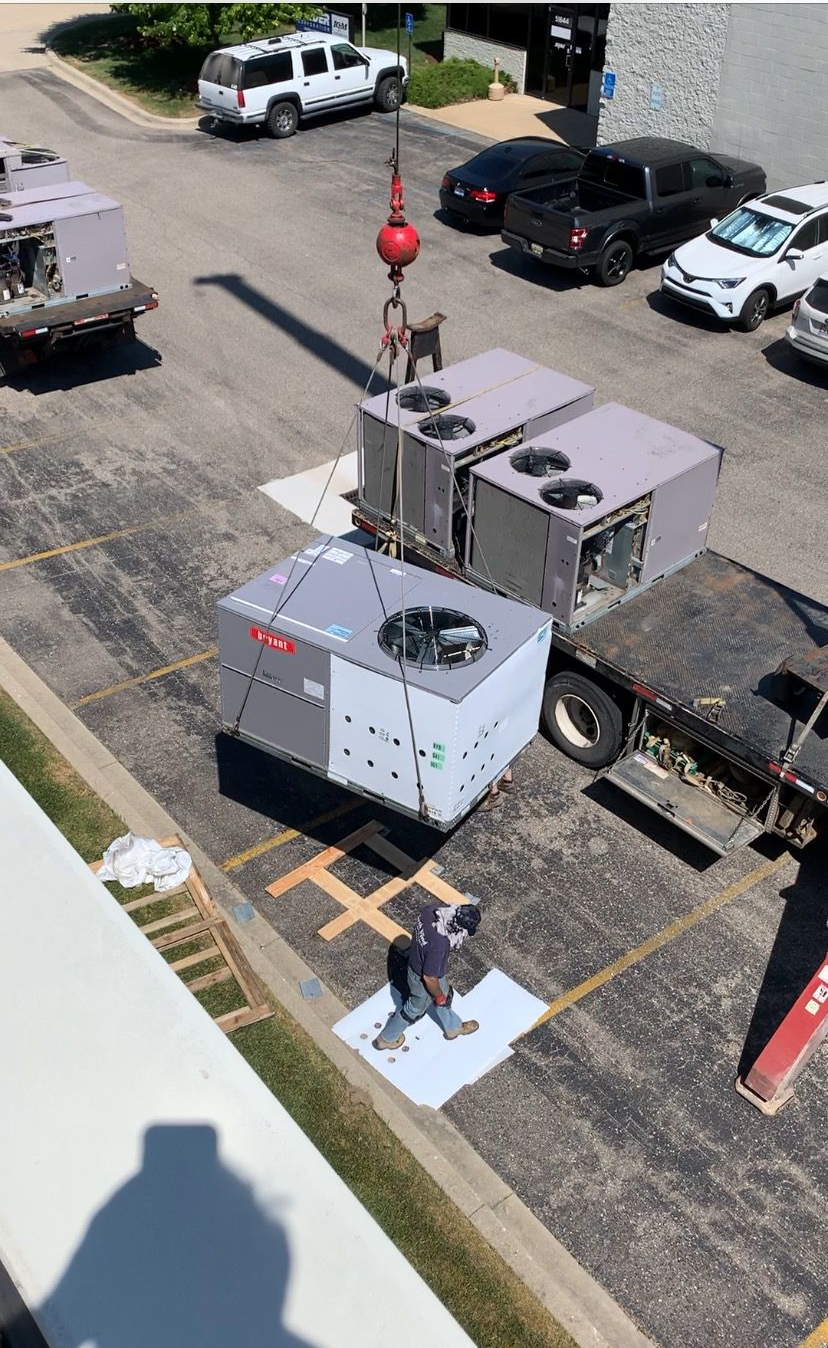 A crane lifting HVAC units in a parking lot. A worker stands on a white square as others stand by.