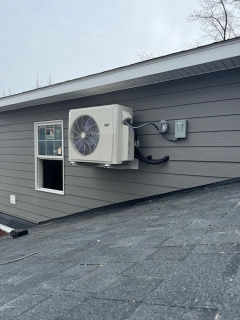 Air conditioner unit mounted on a gray building exterior next to a window. Black pipes extend to a box.