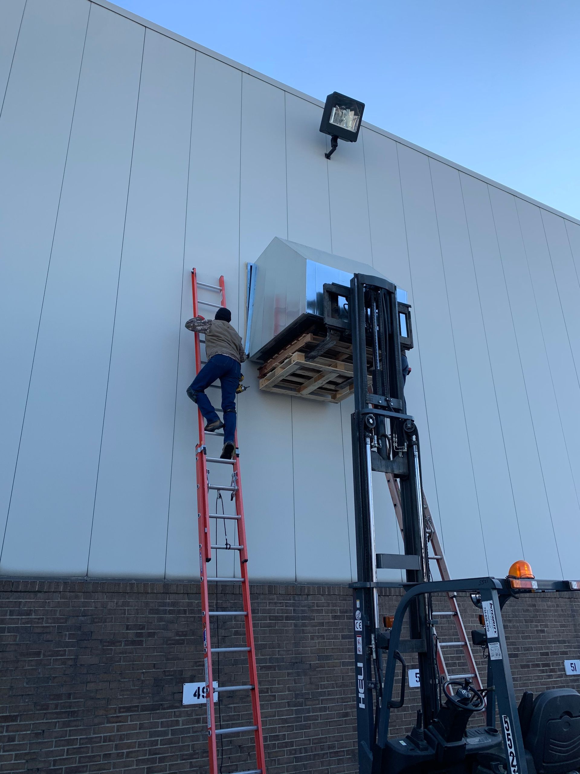 A worker on a ladder assists with a forklift lifting a pallet near a building wall.