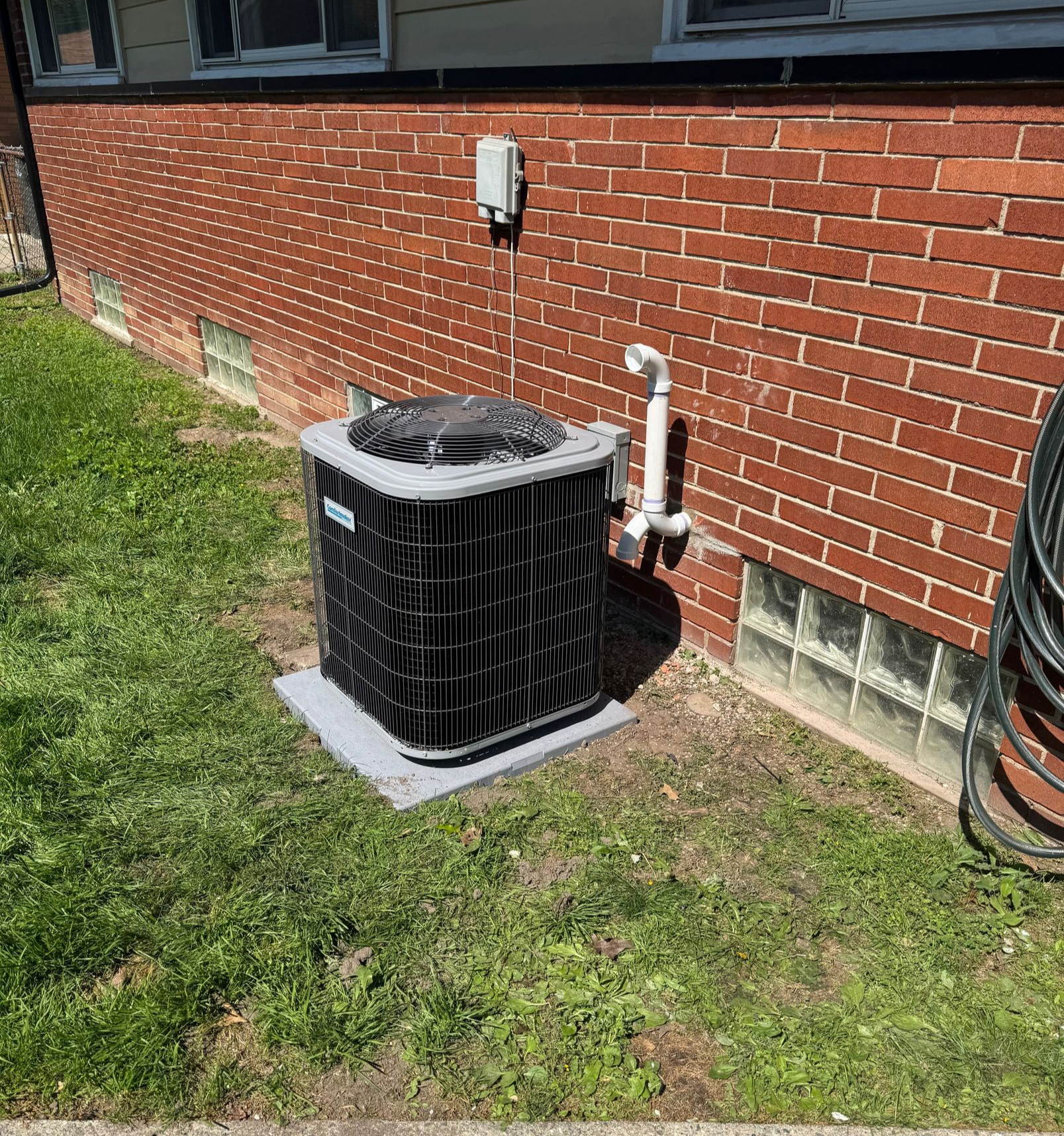 Outdoor air conditioner unit beside a red brick house wall with grass and concrete walkway nearby