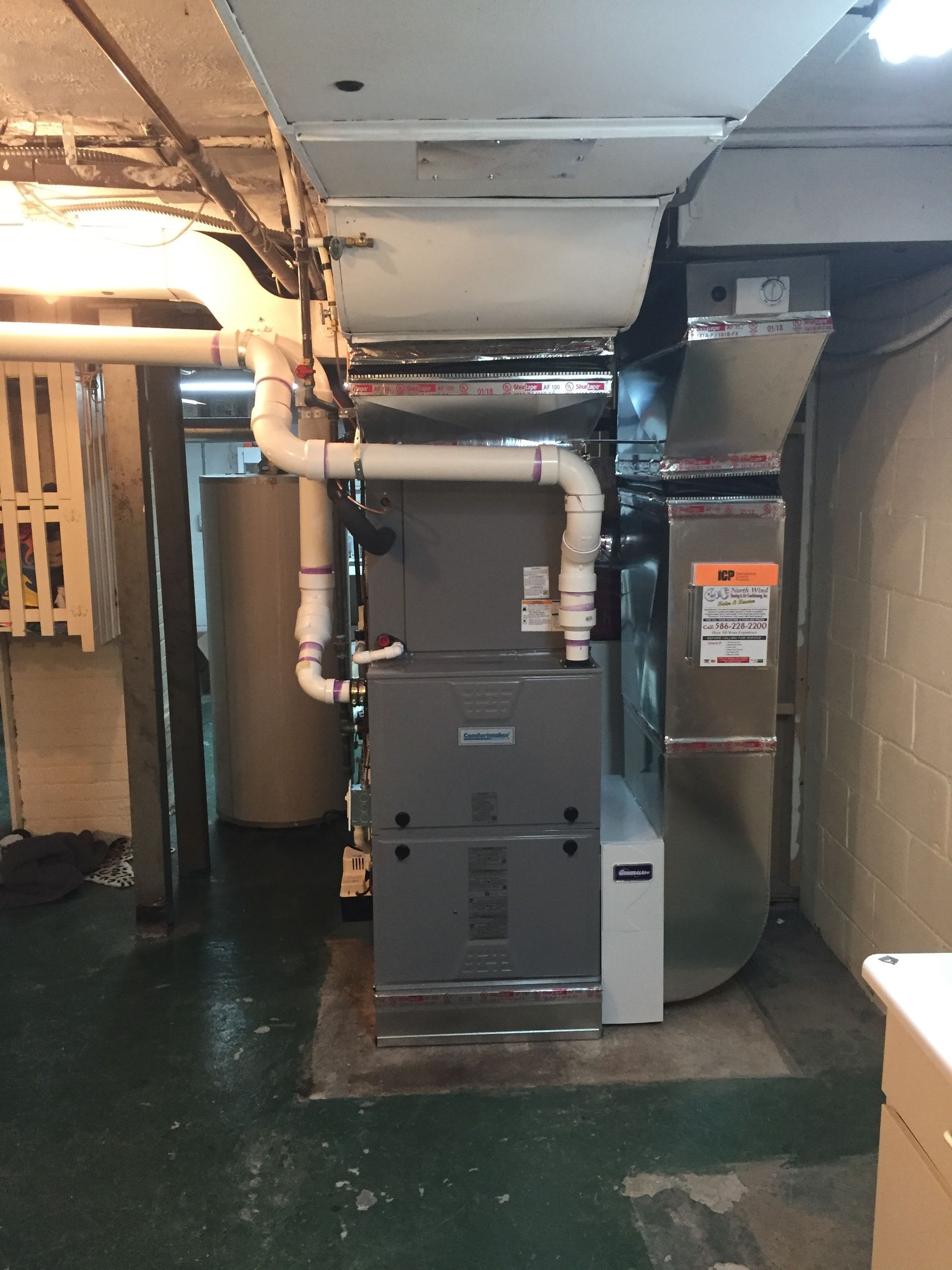 A furnace and ductwork in a basement setting. Gray furnace, white pipes, and silver ductwork.