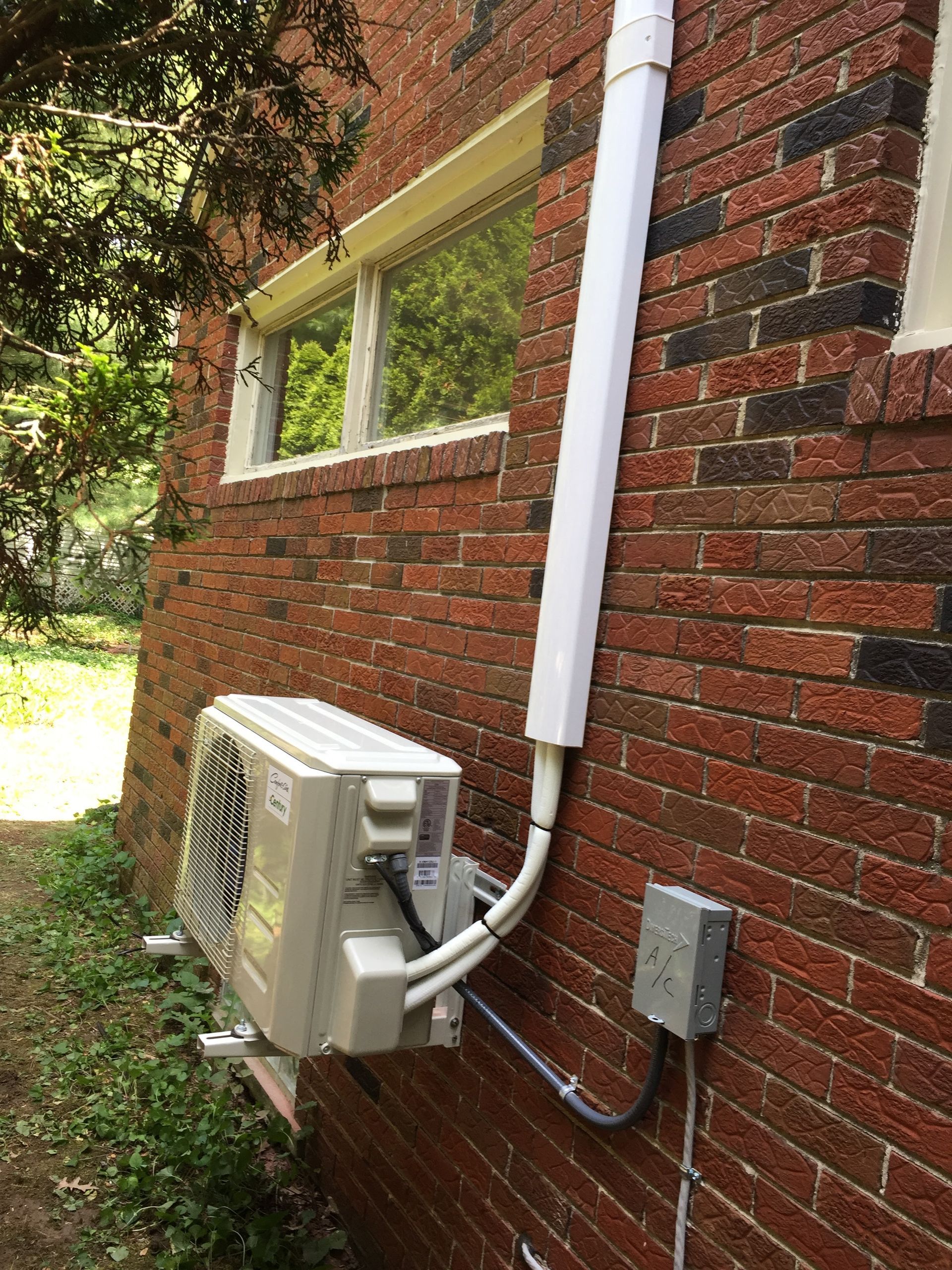 White outdoor AC unit on a brick wall with white conduit and electrical box.