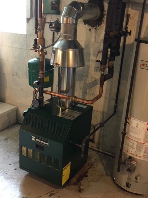 Green boiler with silver vent pipe, copper pipes, and a green expansion tank in a basement setting.