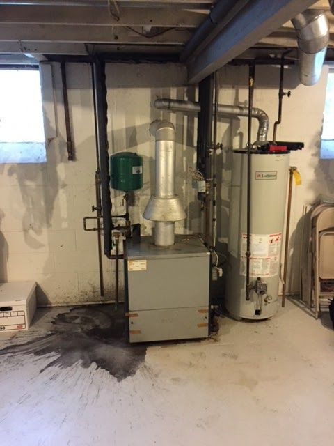 Furnace and water heater in a basement with wet floor. Pipes and a green tank are also visible.