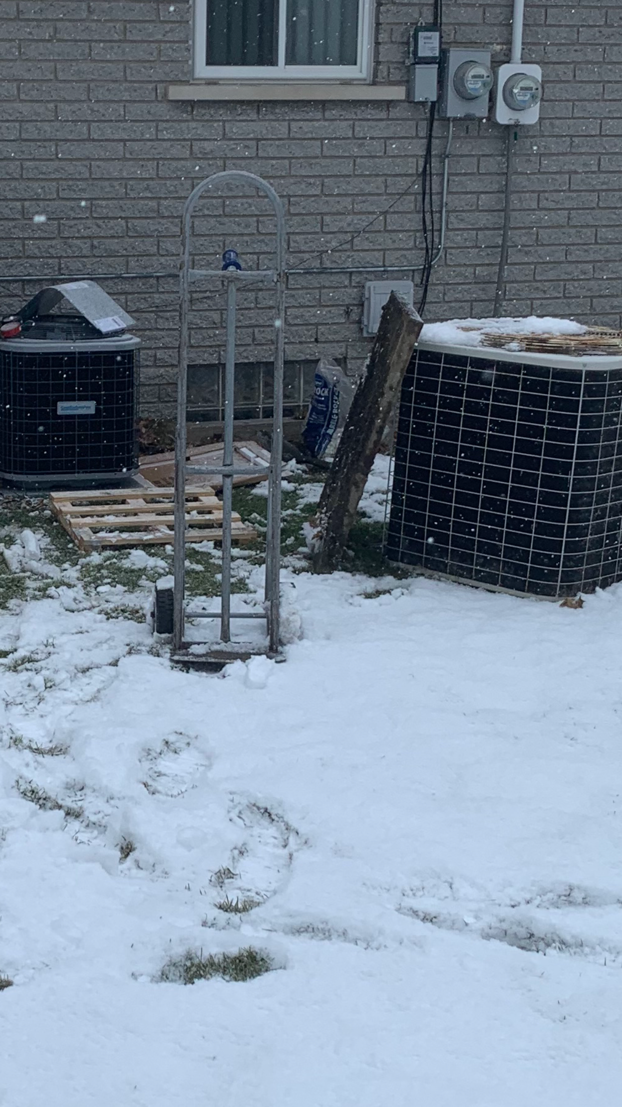Snowy backyard with a hand truck, brick wall, and stacked wood.