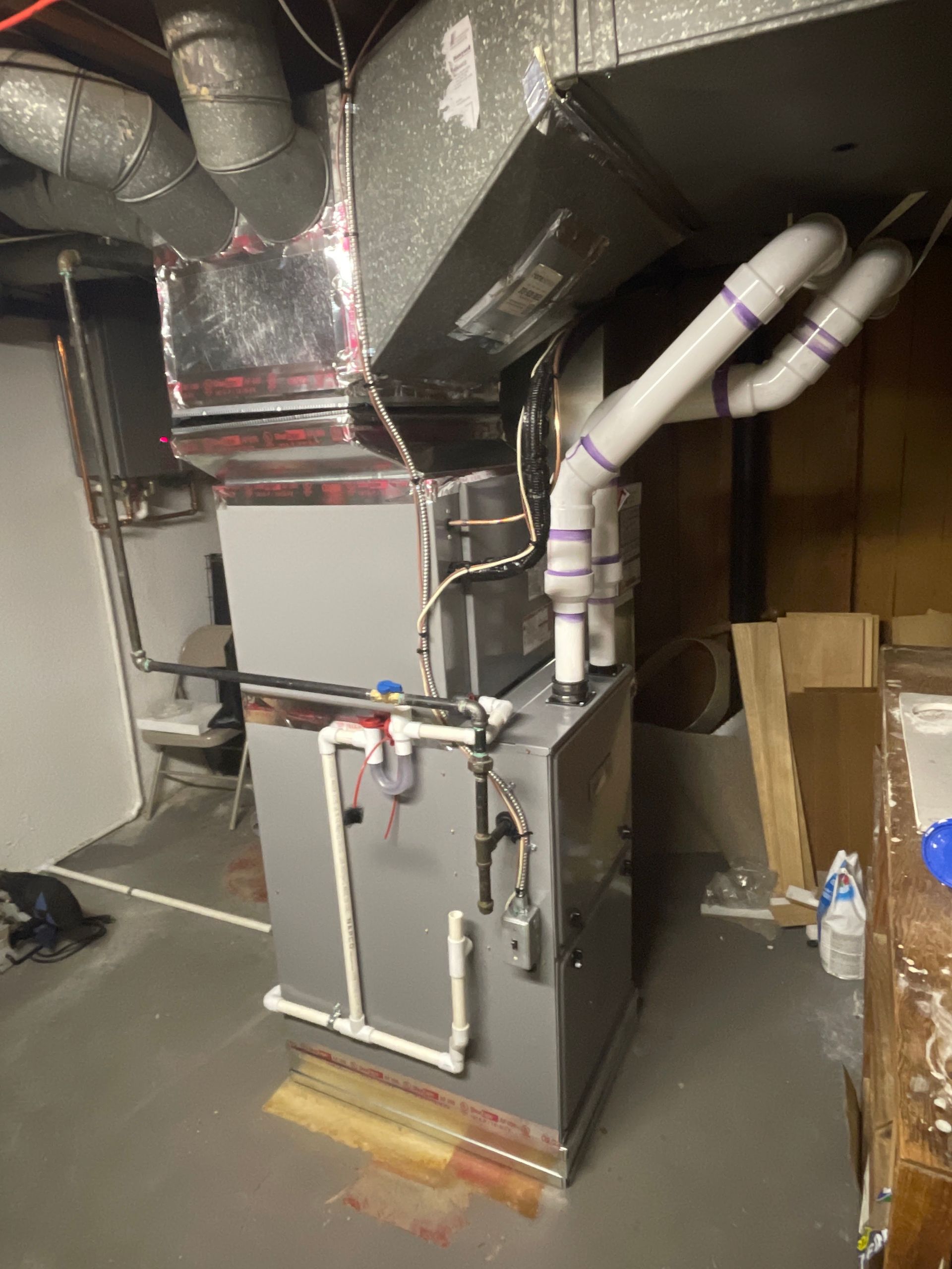 Furnace in a basement with ductwork and PVC pipes. Gray unit with silver ducting and white pipes.