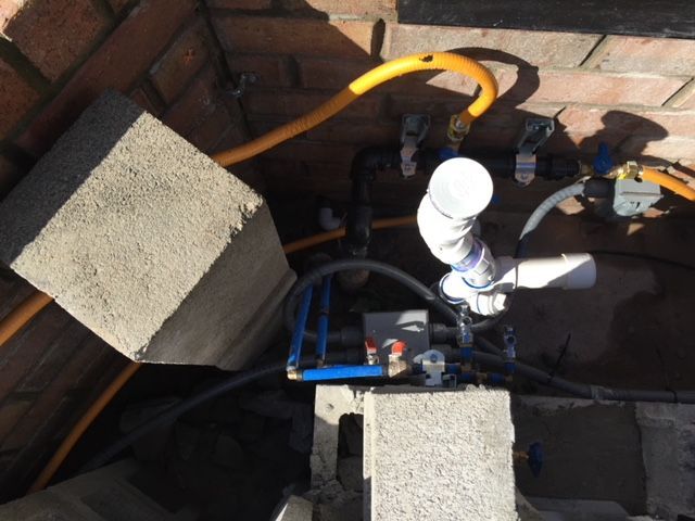 Plumbing with PVC pipes, blue water lines, yellow gas hose, and concrete blocks, against a brick wall.