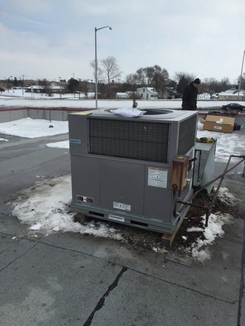 HVAC unit on a snowy rooftop with a person nearby. Gray machinery, overcast sky, snow patches.