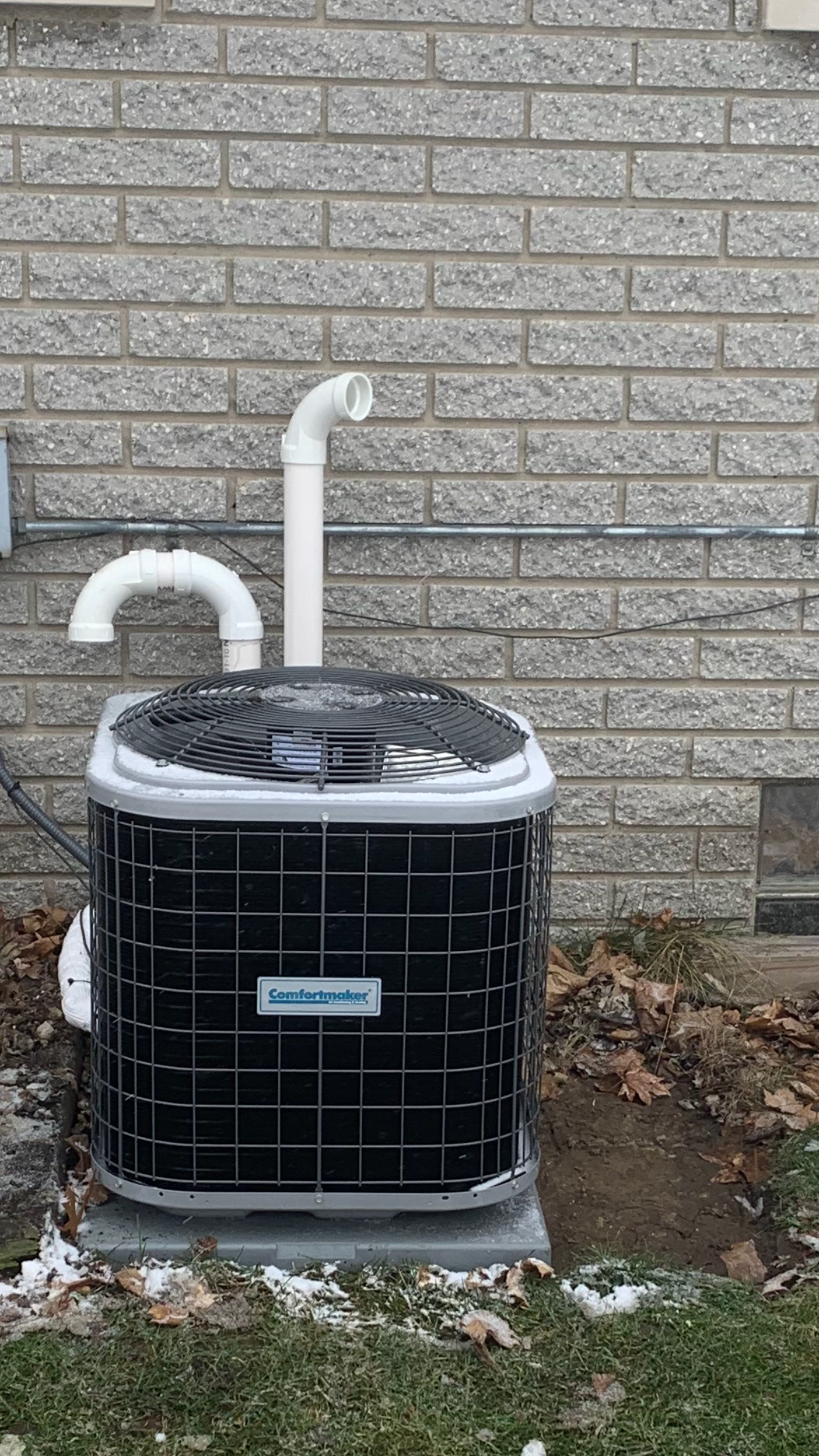 Air conditioner unit against a brick wall, with a white vent pipe.