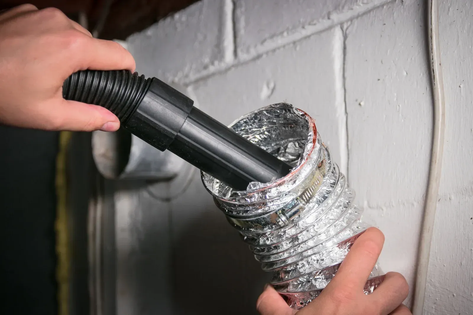 Person using a vacuum hose to clean a dryer vent inside a white wall.