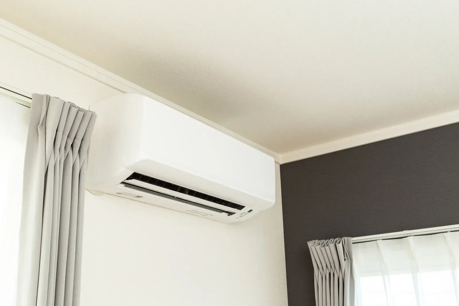 White air conditioner on a wall above a window with gray curtains; interior shot.
