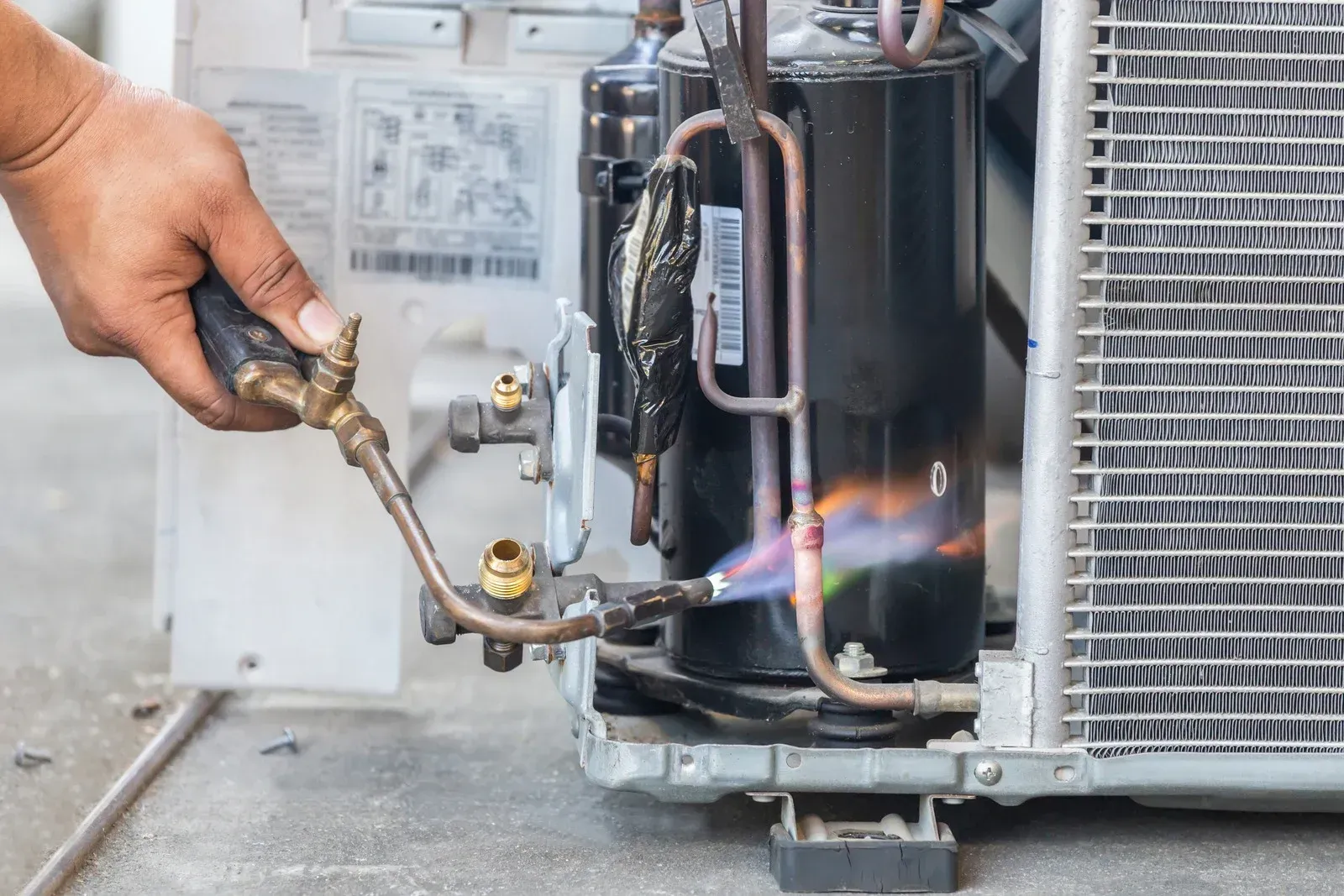 Person using a torch to weld copper pipes on an air conditioning unit; flames are visible.