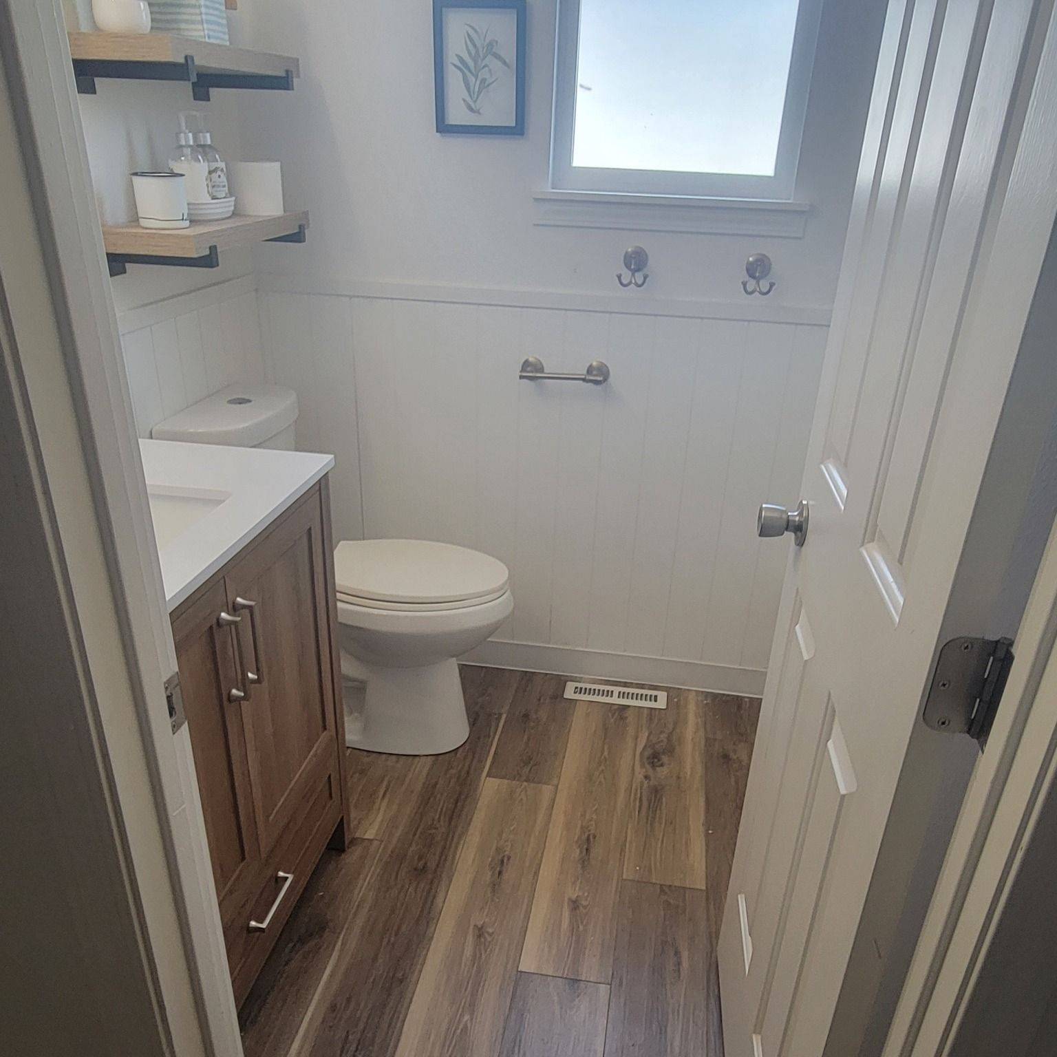 Bathroom with wood-look floor, white toilet, wood vanity, shelves, and white door.