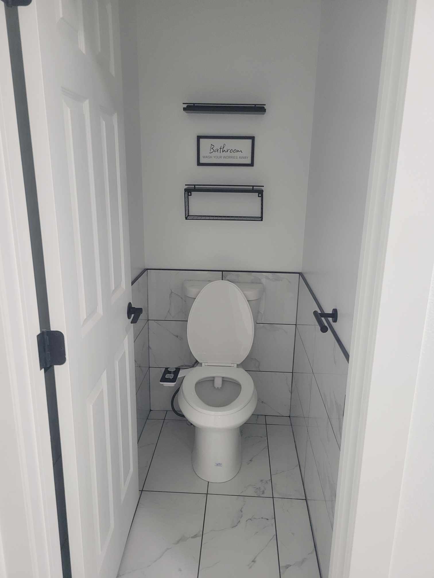 A small, white, tiled bathroom with a toilet, shelves, and an open door; black accents.