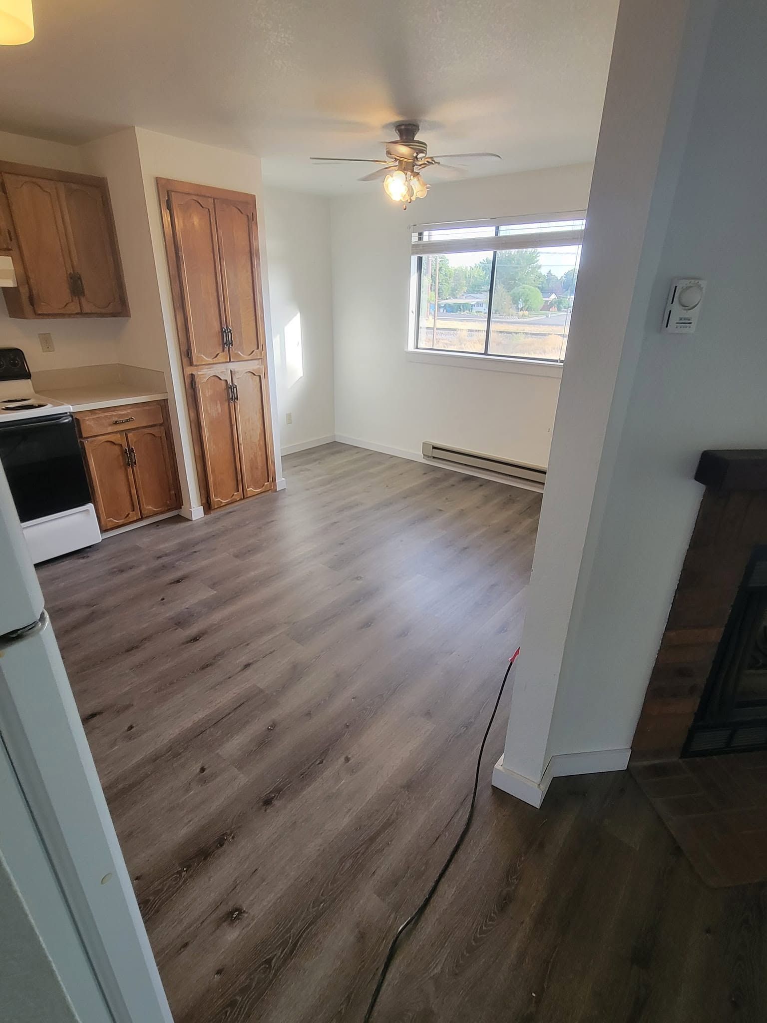 Empty kitchen and dining area with wood floors, white walls, ceiling fan, and daylight from a window