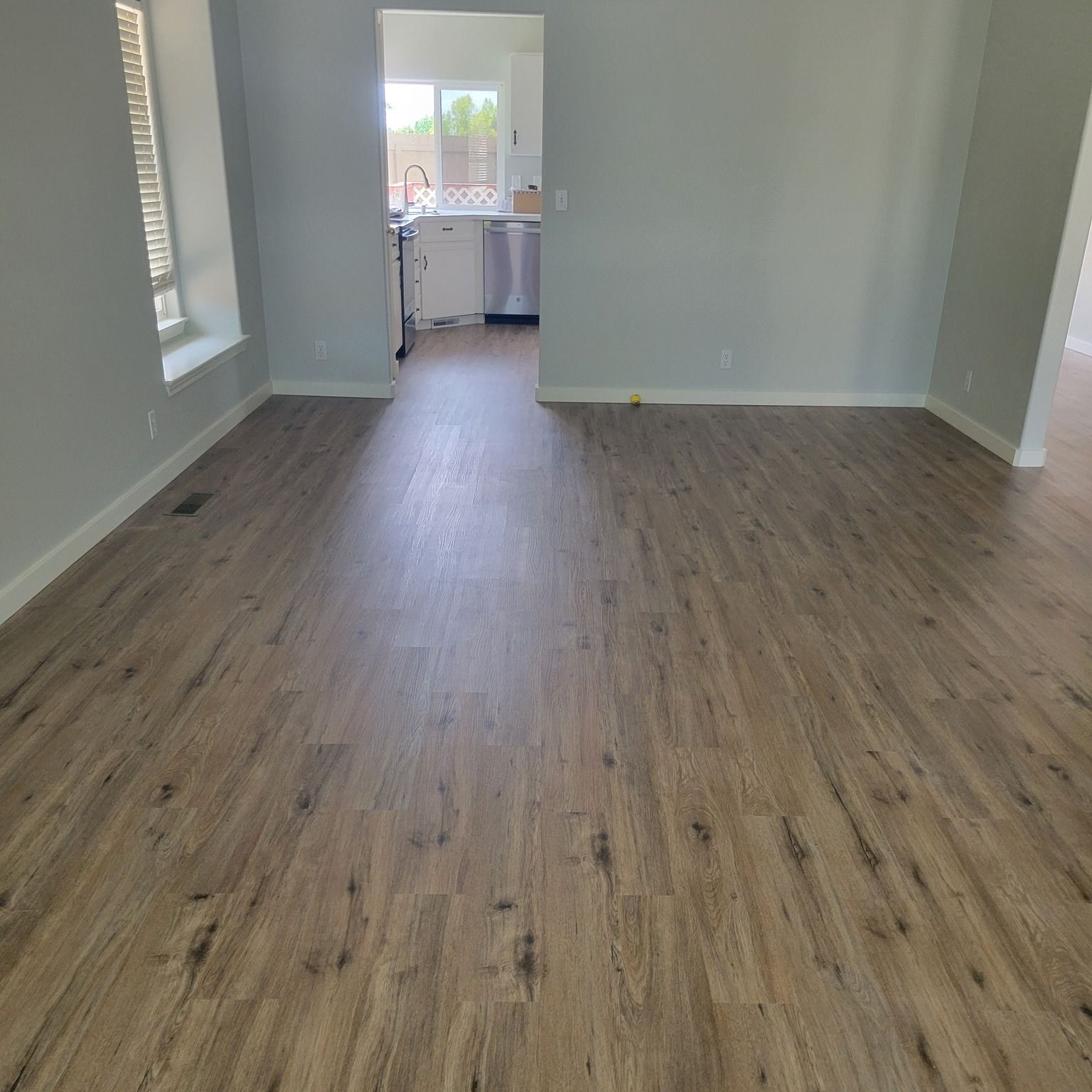 Empty room with wood-look laminate flooring and light gray walls. Kitchen visible in the background.