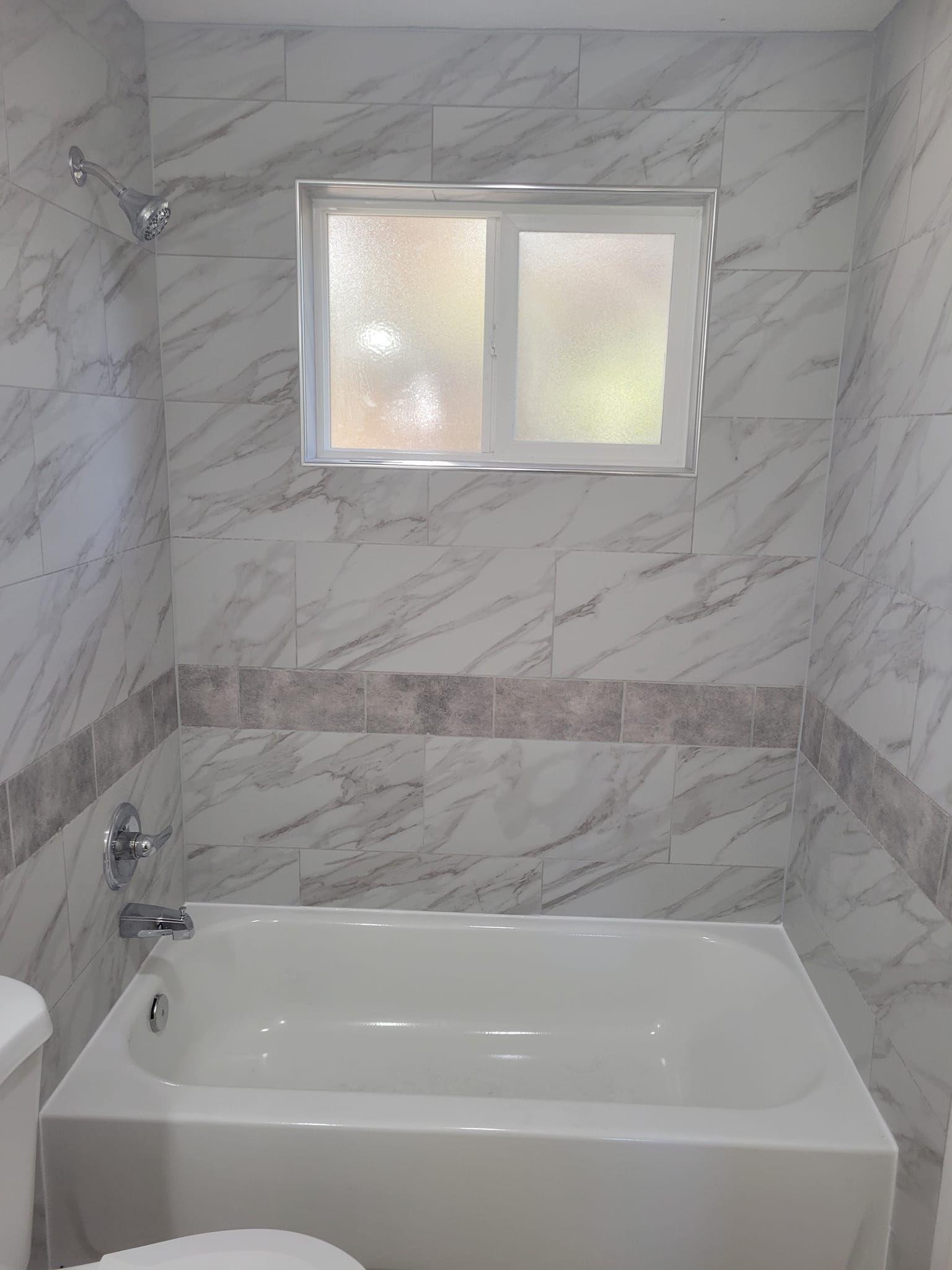 Bathroom with white tub and marble-look tile; a window, showerhead, and silver fixtures.