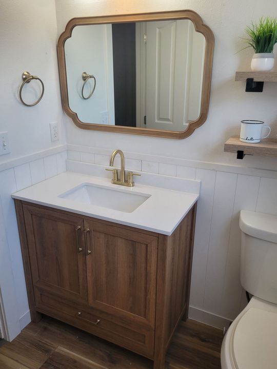 Bathroom with wooden vanity, gold faucet, white countertop, and scalloped mirror.