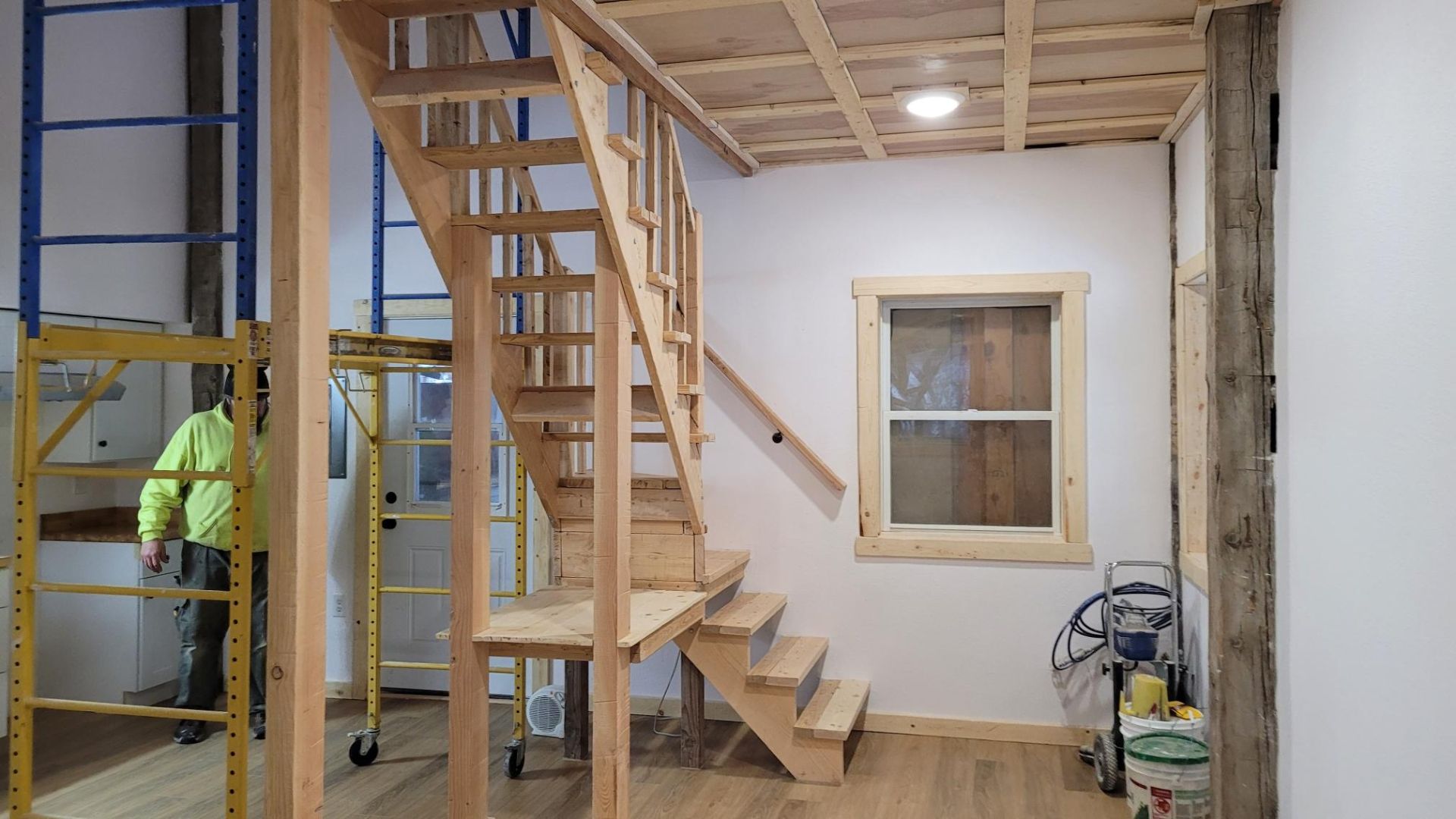 Interior of a building under construction, showing wooden staircase, window, and a person in the background.