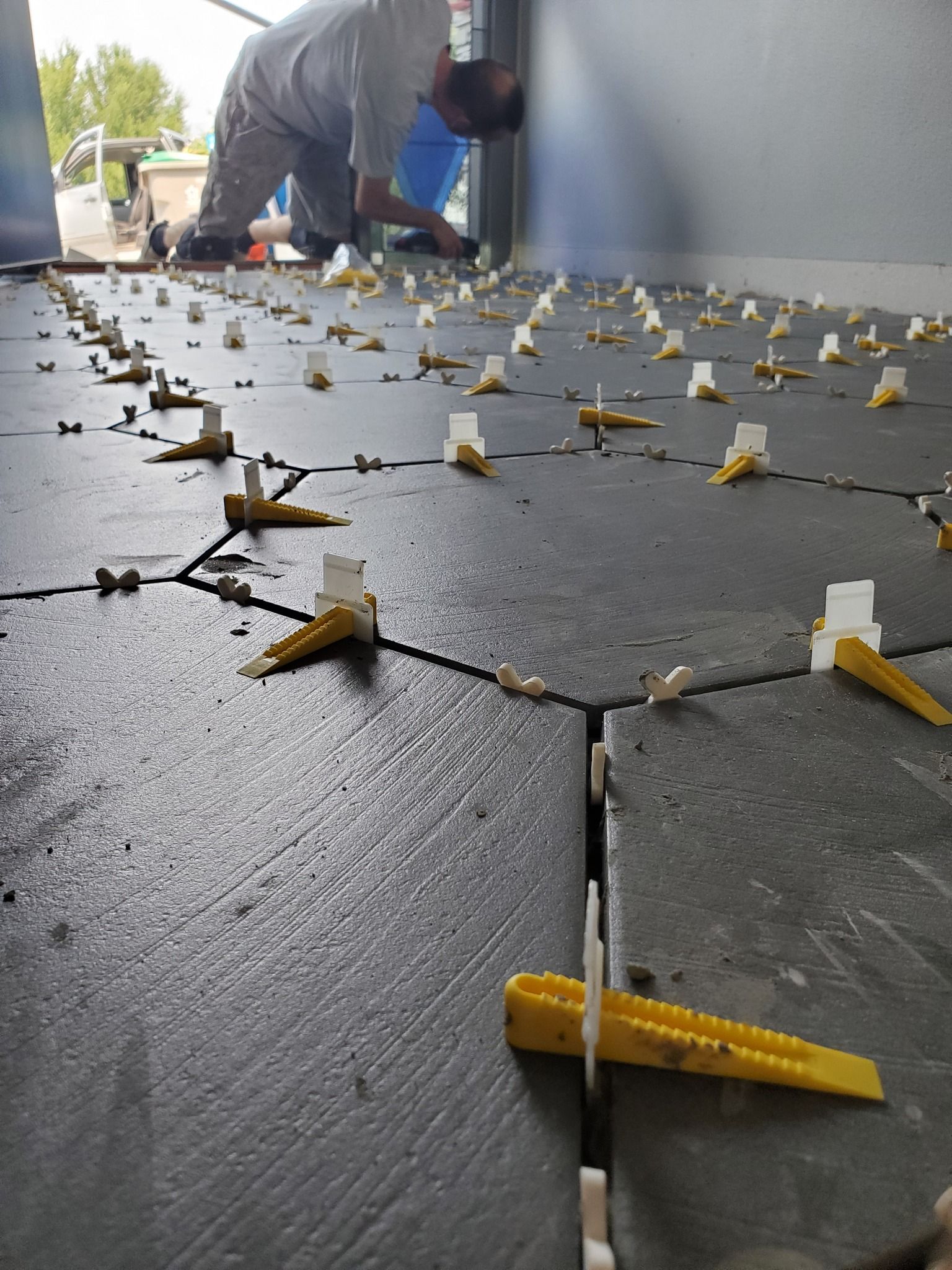 Person installing dark floor tiles, using leveling clips. Interior setting with open doorway.