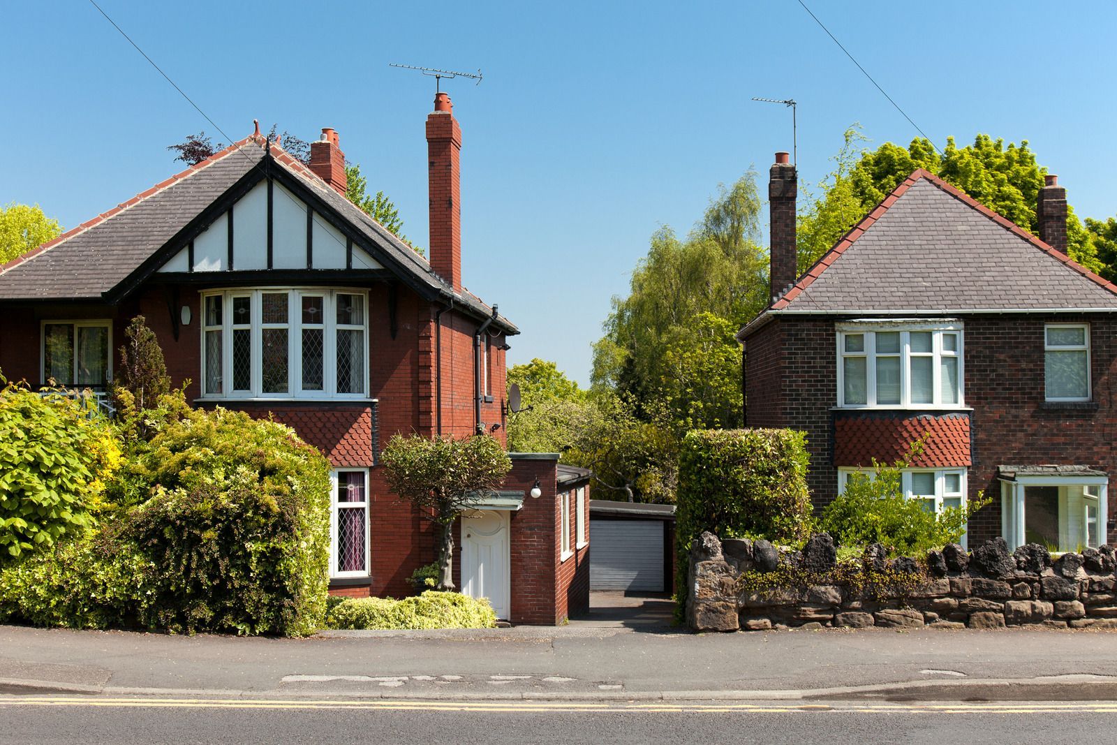 Two suburban houses with brick facades, front gardens, and a road in the foreground.