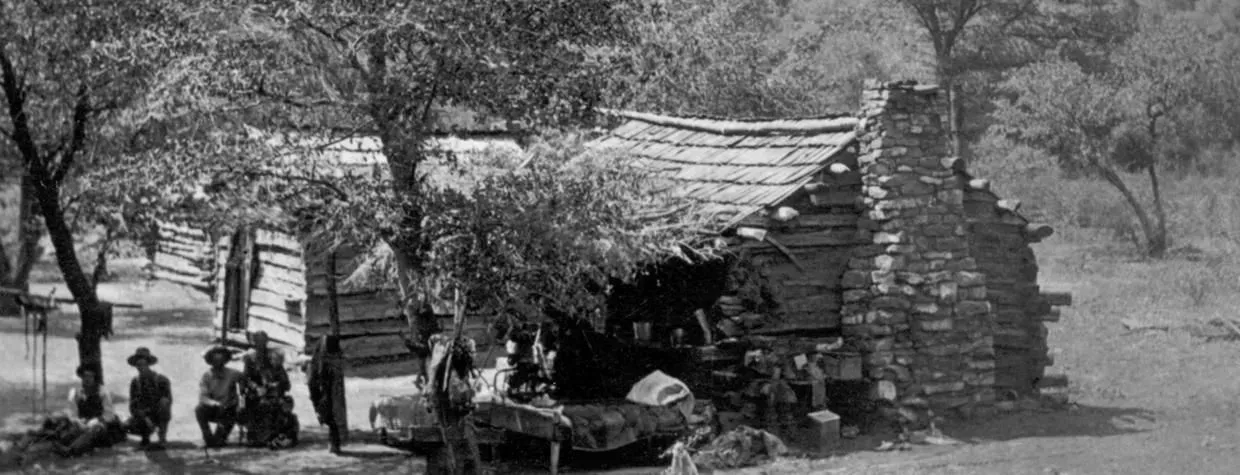 People gathered outside a rustic stone cabin in a wooded rural setting.