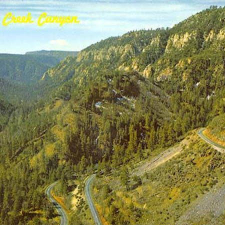 Mountain road winding through a forested canyon with steep rocky cliffs and yellow script text at top left