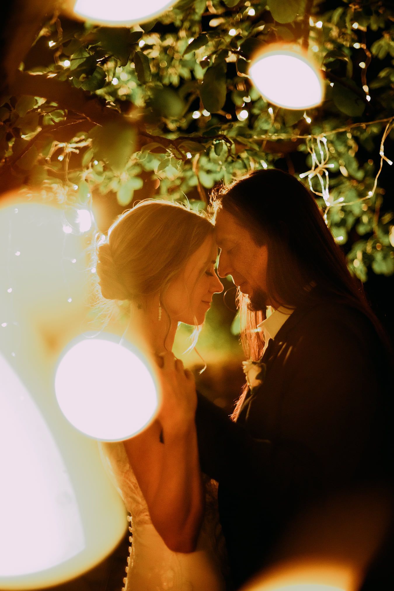Two people touching foreheads under warm string lights, silhouetted in a romantic outdoor setting