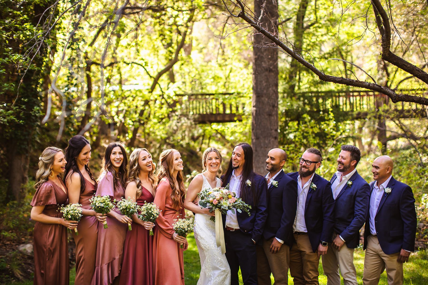 Wedding party in formal attire poses outdoors among trees, with the bride holding a bouquet.