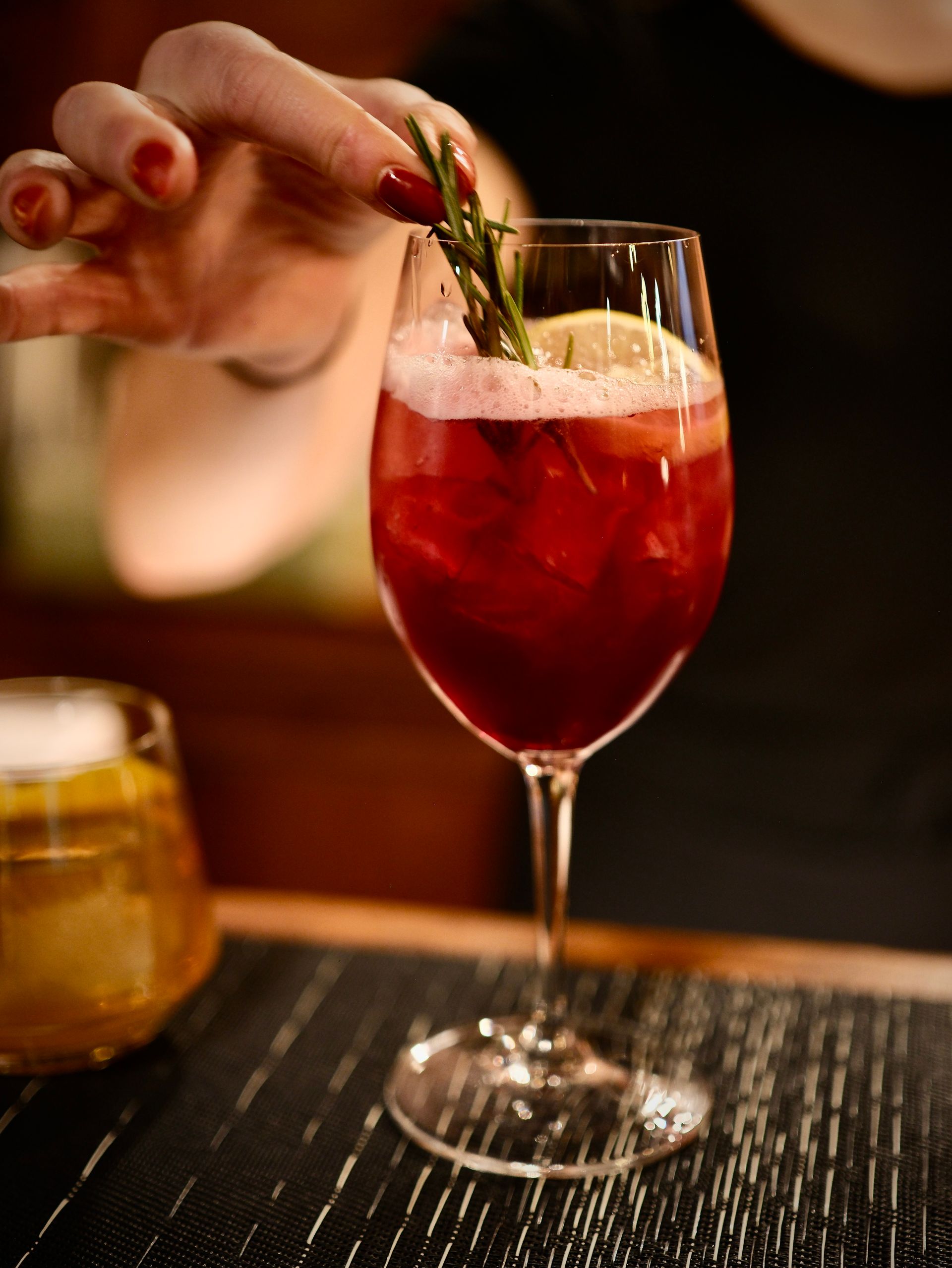 Hand placing a mint sprig in a red cocktail glass on a bar counter