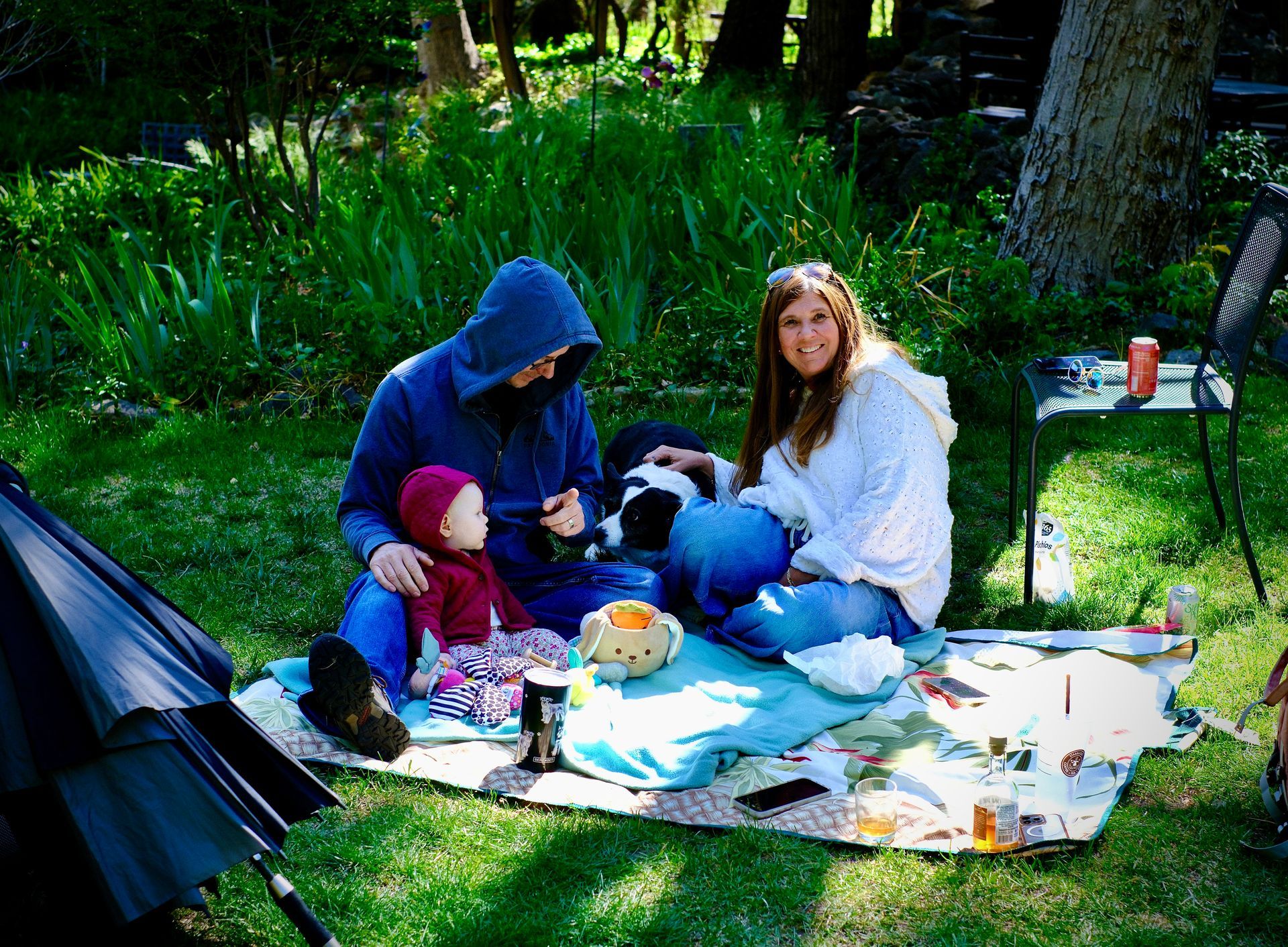 Two people sitting on a picnic blanket in a grassy park, surrounded by bags, bowls, and camping gear.