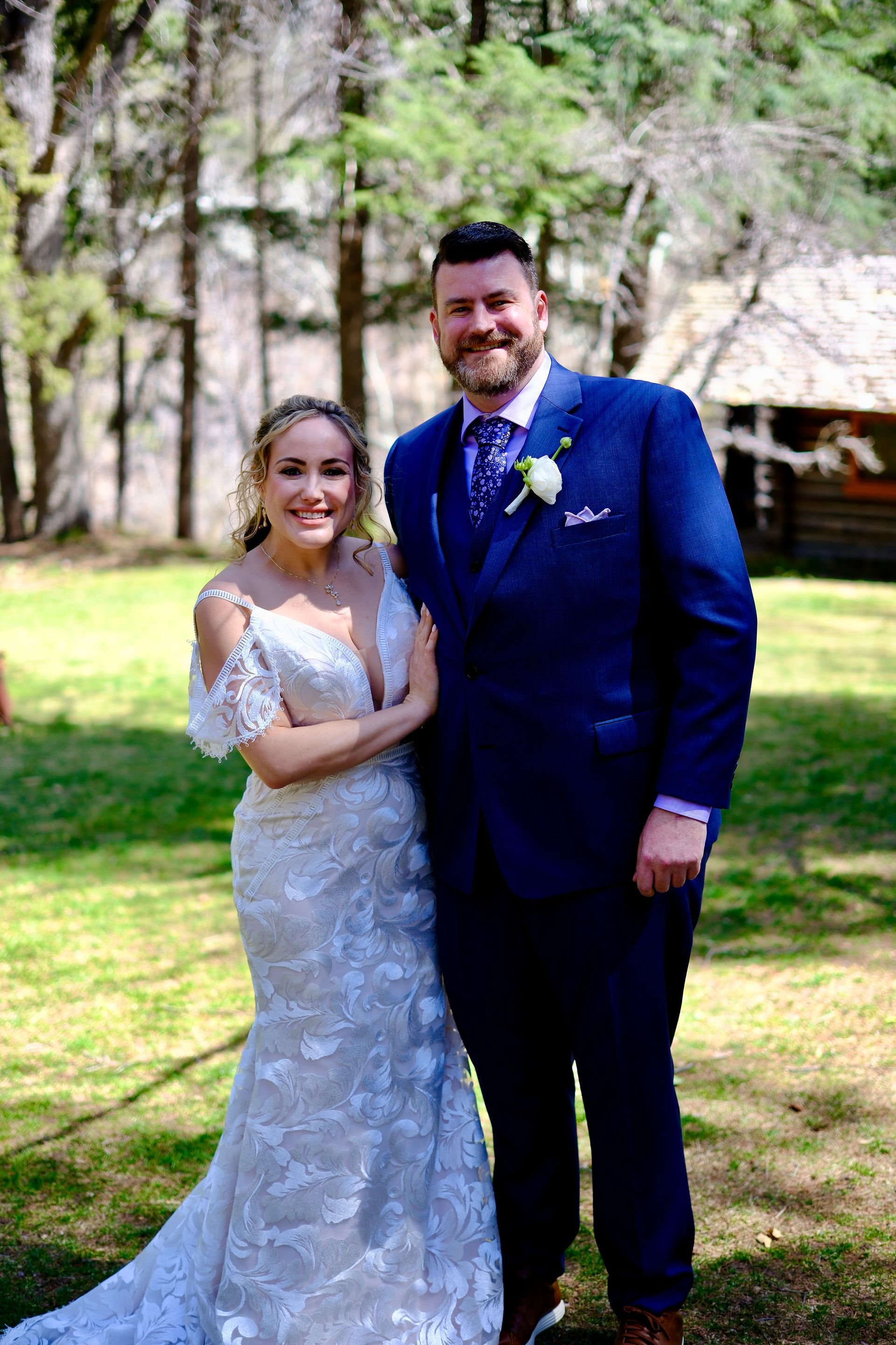 Bride and groom posing outdoors, smiling in wedding attire, with trees and grass in the background.